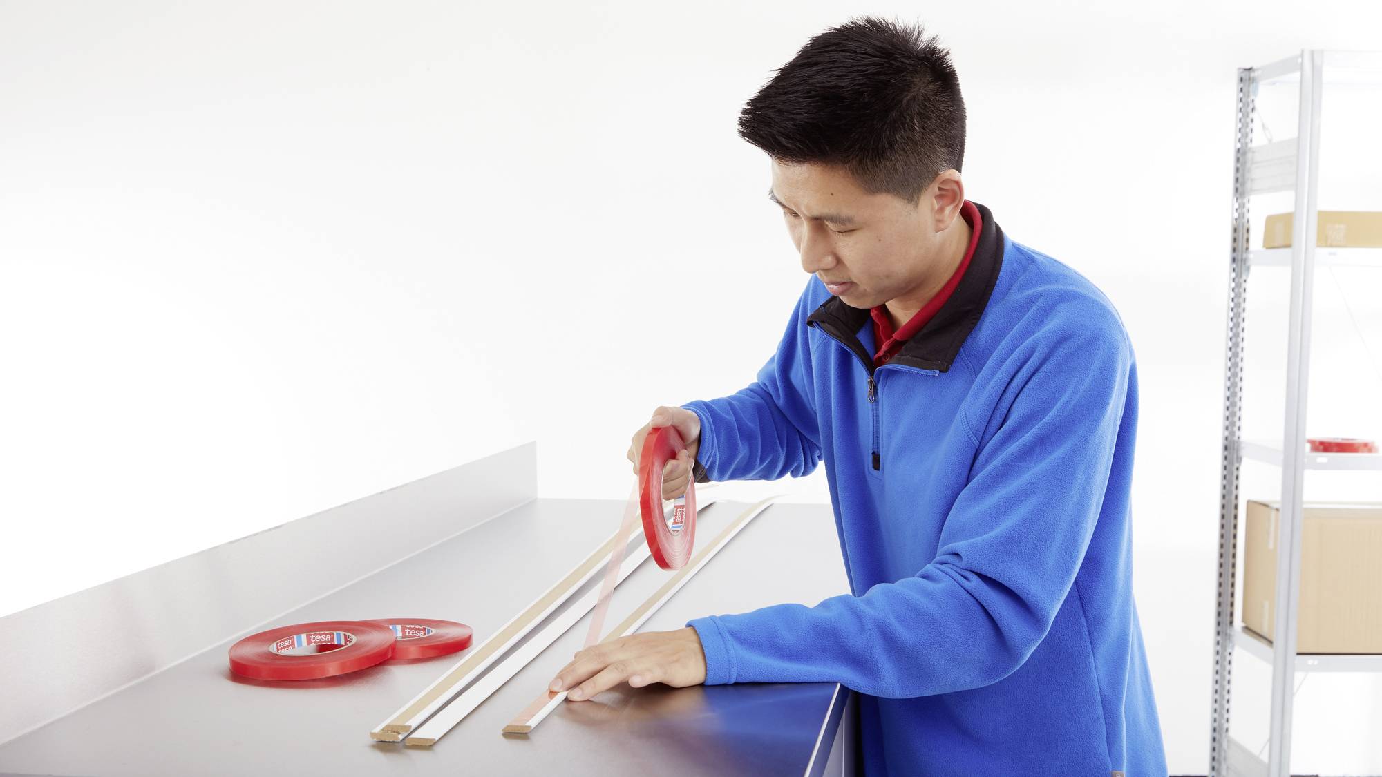 A man in a blue jumper is cutting insulating tape at a workbench. Shelving and boxes are visible in the background.