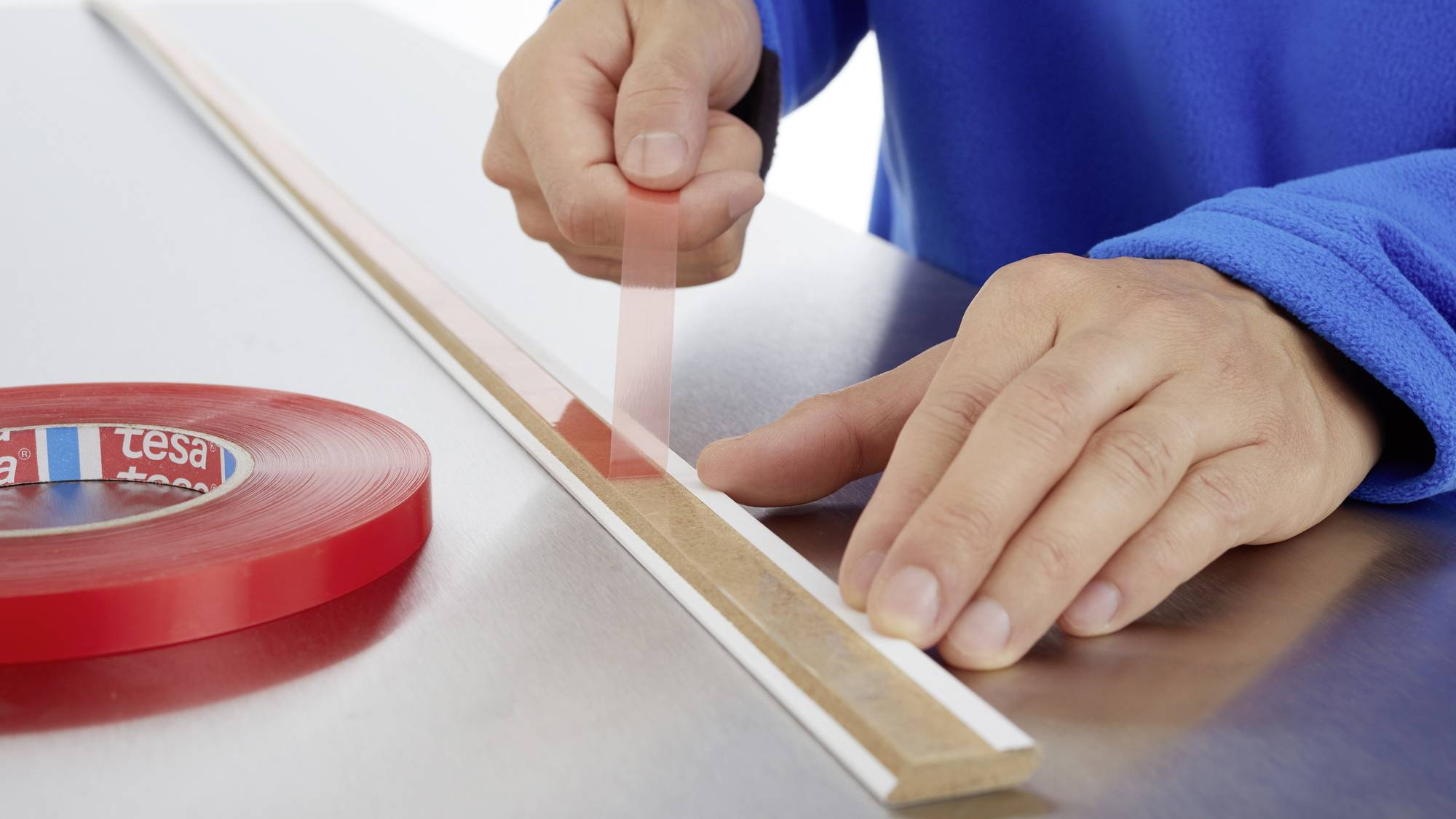 A person is sticking a long, narrow piece of wood onto a table using red double-sided tape.