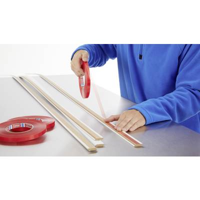 A person wearing blue clothing is applying double-sided tape to a wooden batten, with several battens and tape rolls lying nearby.