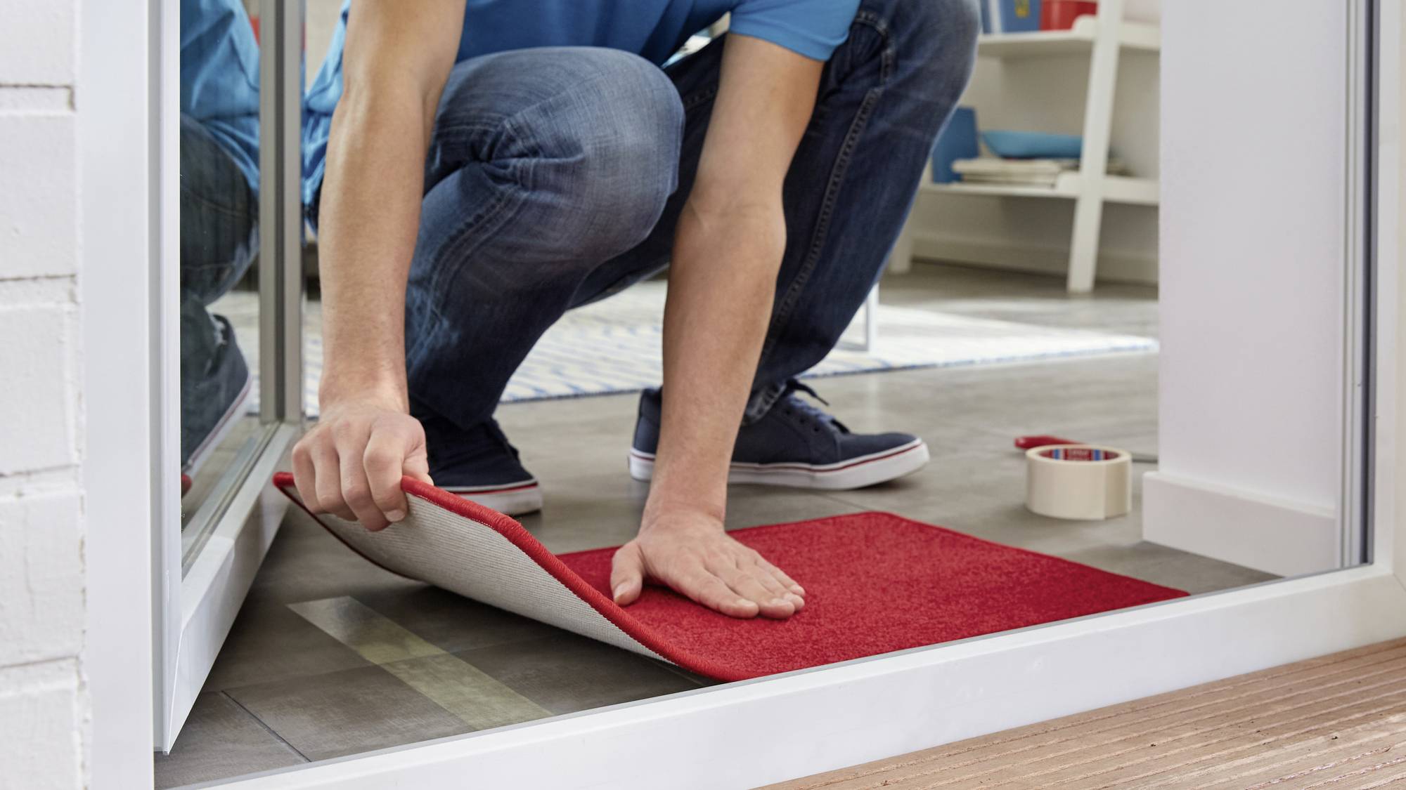 A person is laying a red carpet runner in a room. A roll of double-sided tape and a pair of scissors are placed nearby.