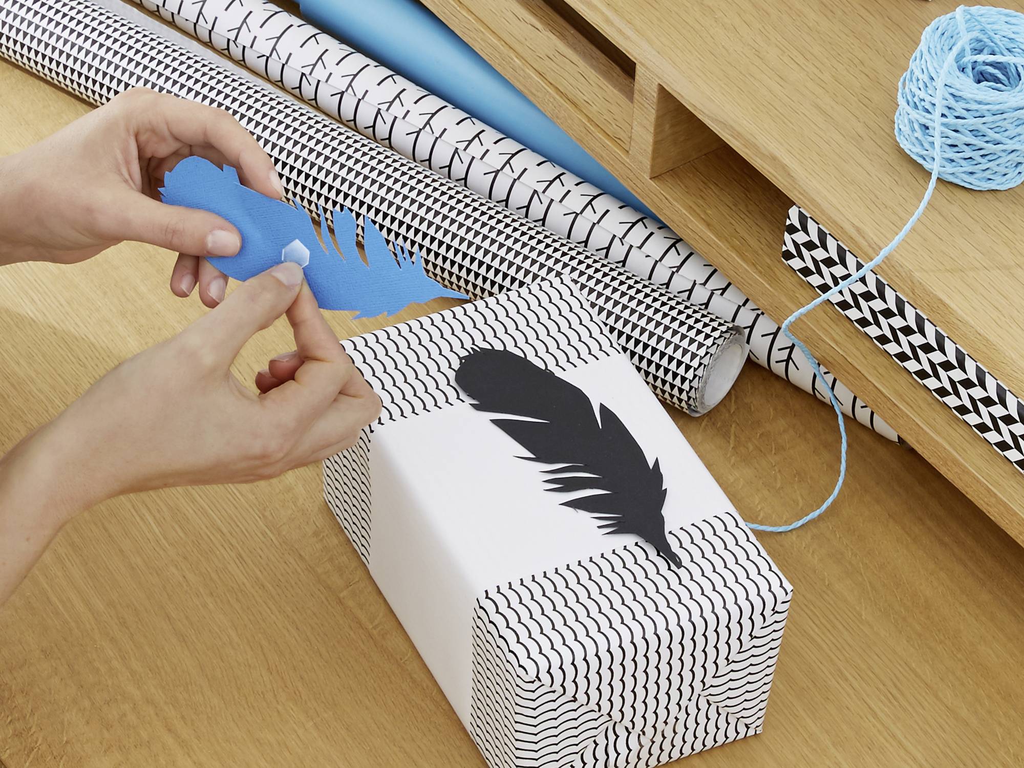 A person is decorating a gift with blue paper and a black feather. The gift is wrapped in white and black patterned paper.