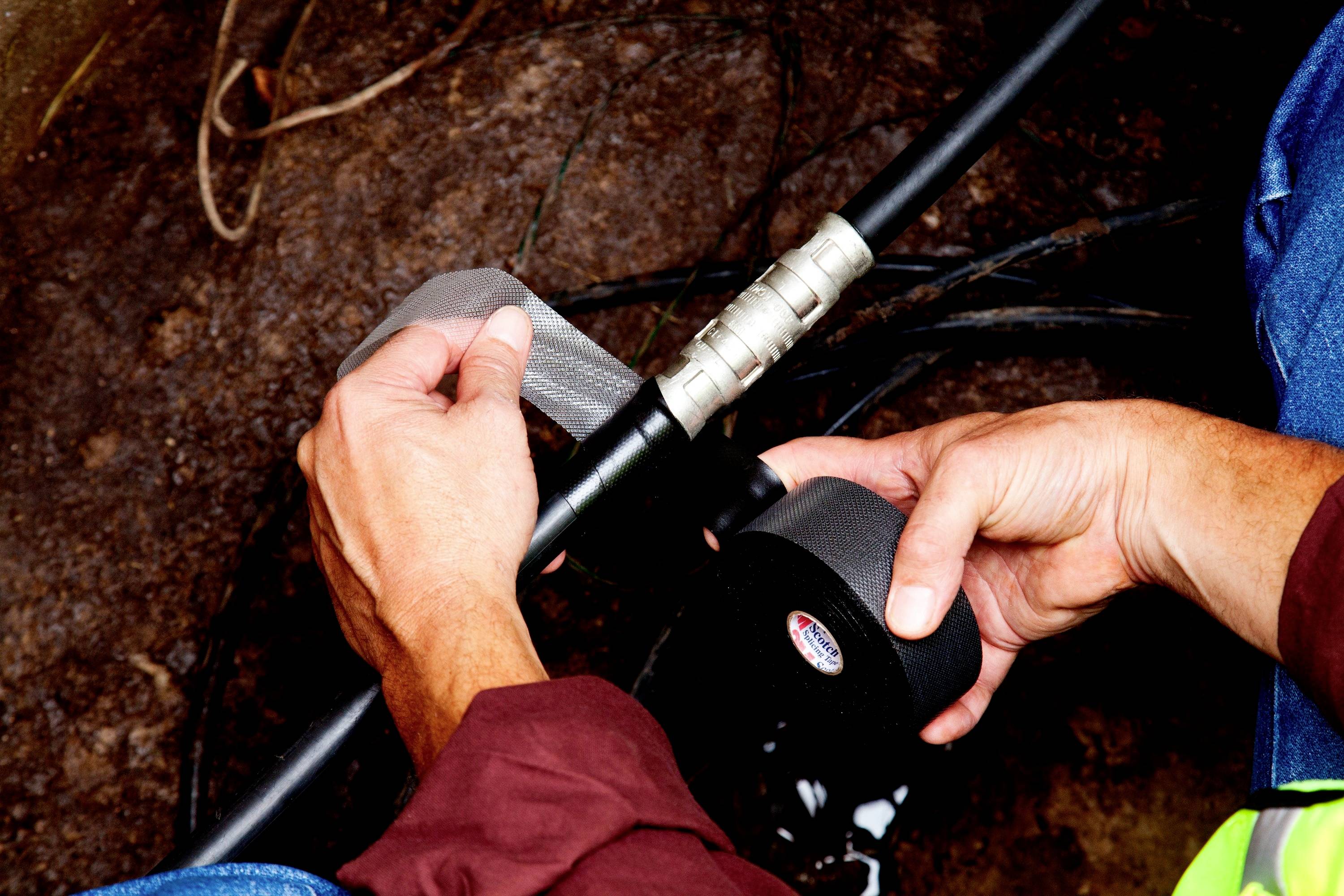 'Person Wrapping Cable with Insulating Tape' - Hands are wrapping two types of insulating tape around a thick black cable. The environment appears industrial or workshop-like.