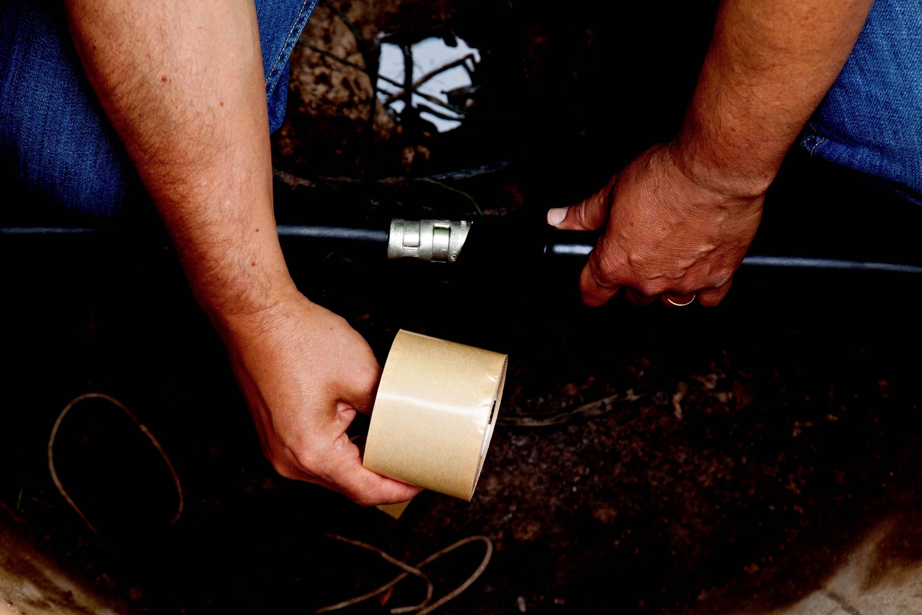 A man is repairing a water pipe outdoors using adhesive tape. Hands are holding the pipe and tape; ground moisture is visible in the background.
