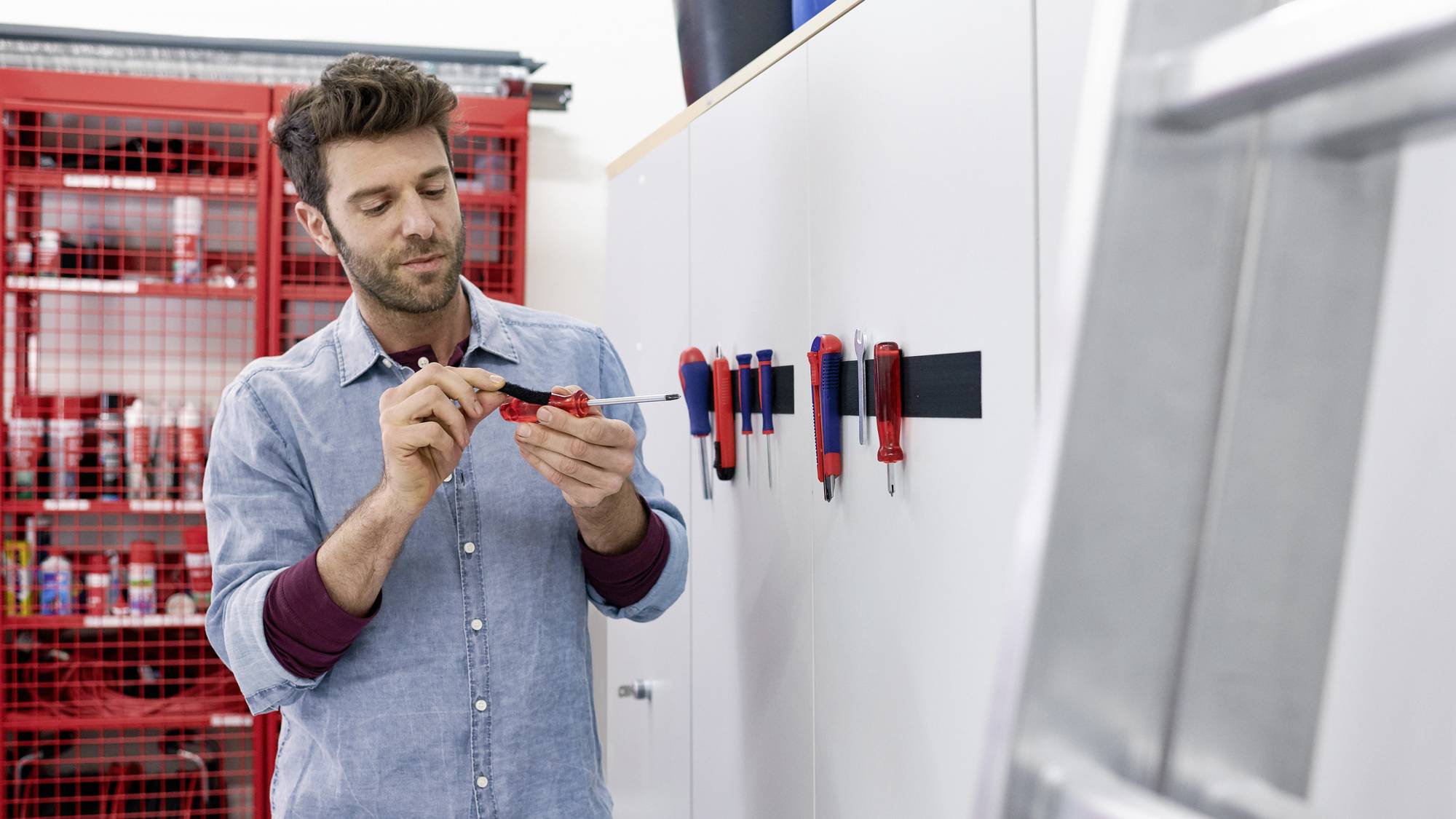 A man is repairing a small device with a screwdriver in a workshop with red shelves full of tools in the background.