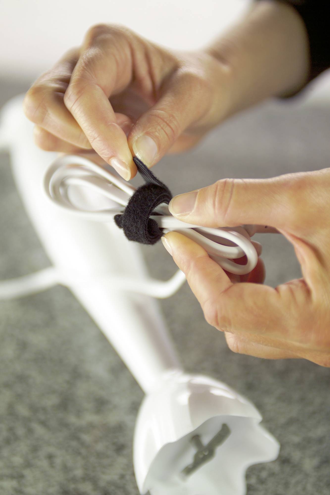 A person is fastening the cable of a white hand mixer together with a black cable tie.