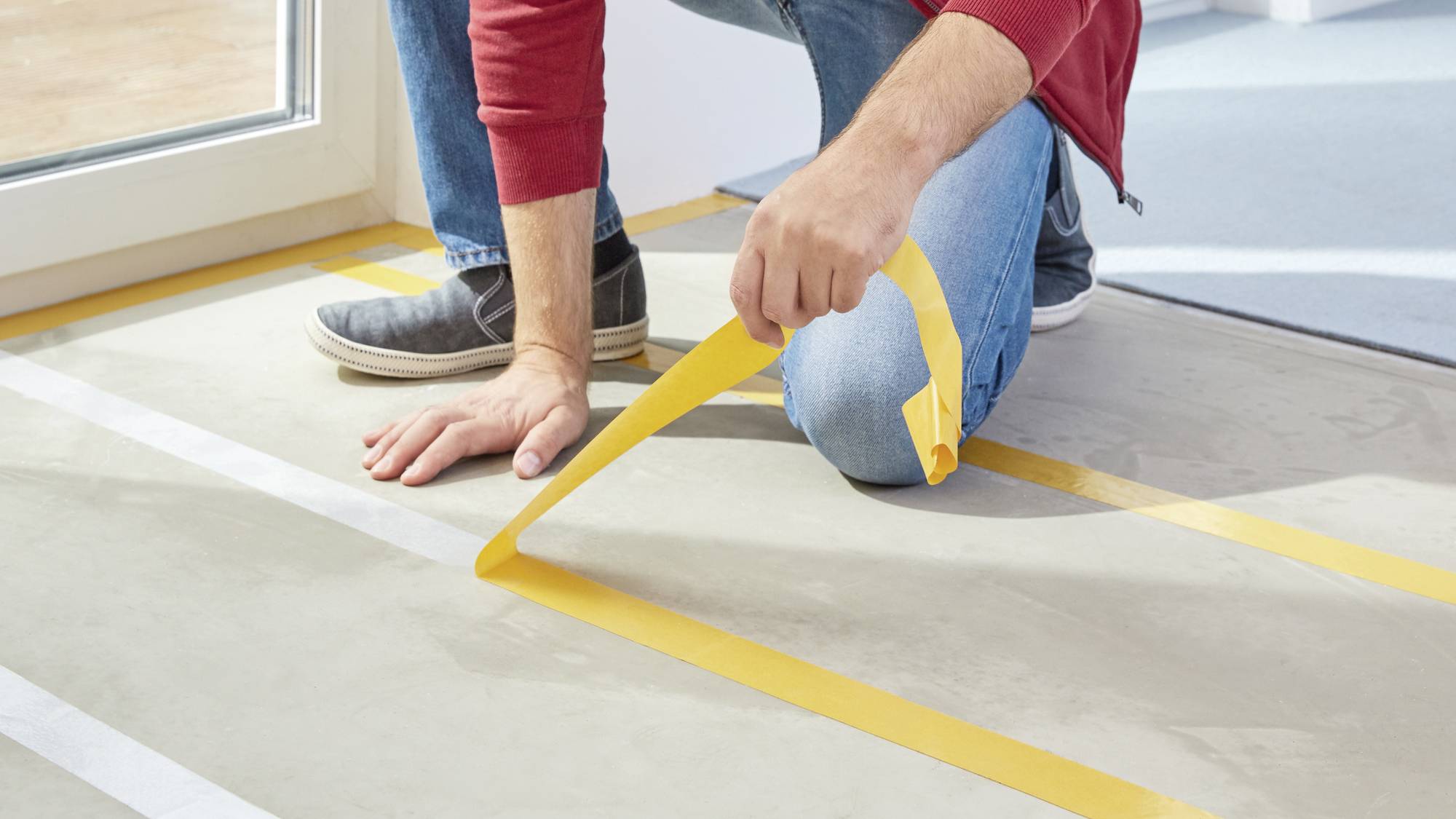 A person is kneeling on the ground and sticking yellow tape in strips onto a light-coloured floor, likely for marking or construction purposes.
