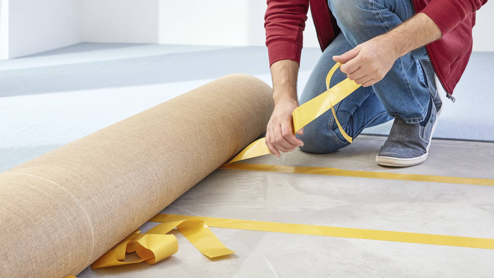 A person is laying carpet by using yellow adhesive tape. Hands are securing the tape to the floor, carpet is being unrolled.