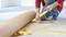 A person is laying carpet by using yellow adhesive tape. Hands are securing the tape to the floor, carpet is being unrolled.