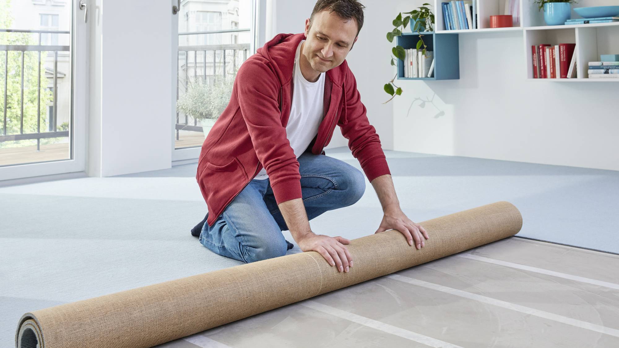 A man in a red hoodie is kneeling on the floor in a bright room, unrolling a carpet. Shelves and windows are visible in the background.