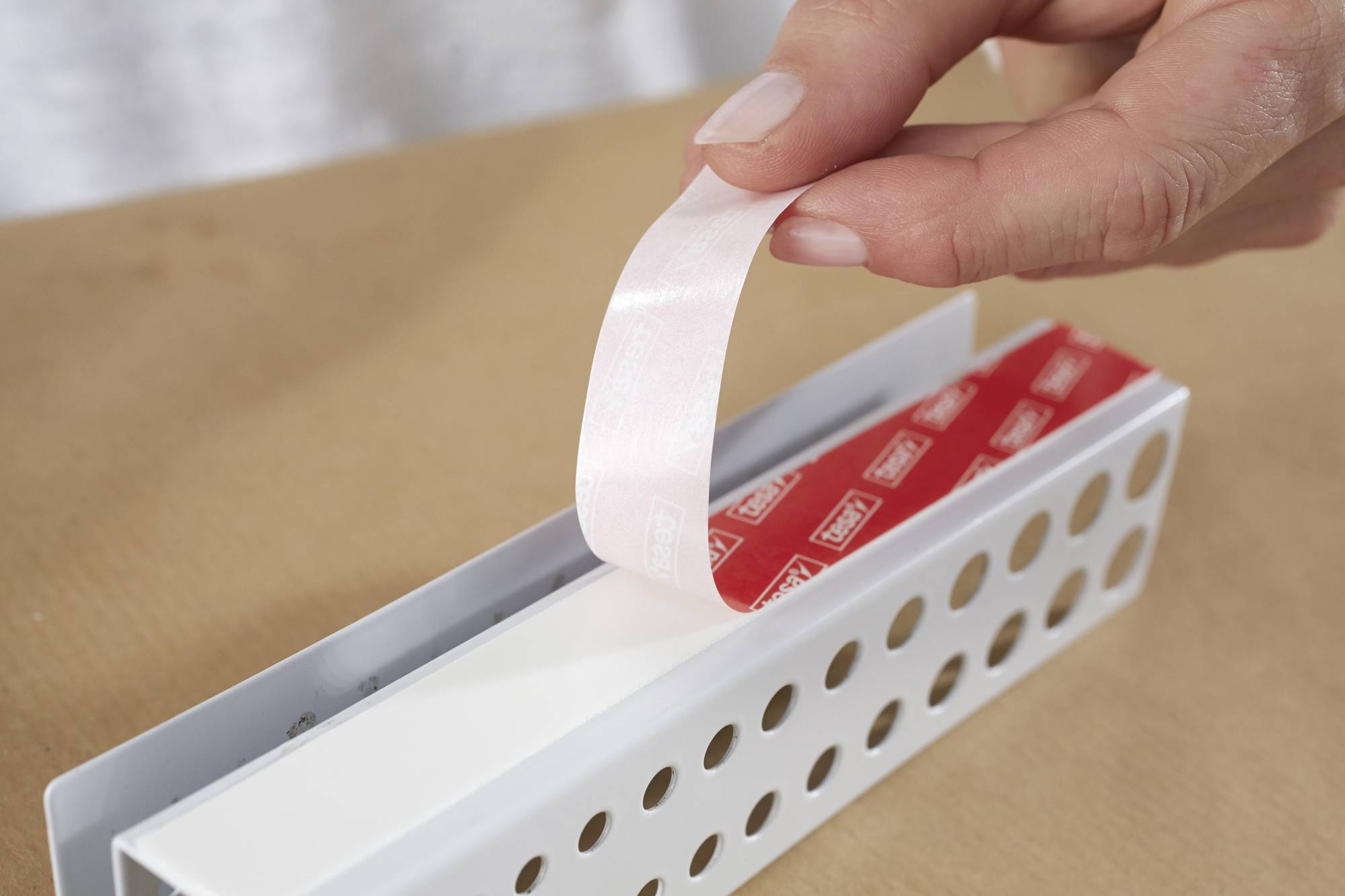 A hand is peeling a double-sided adhesive tape from a perforated metallic tray lying on a table.