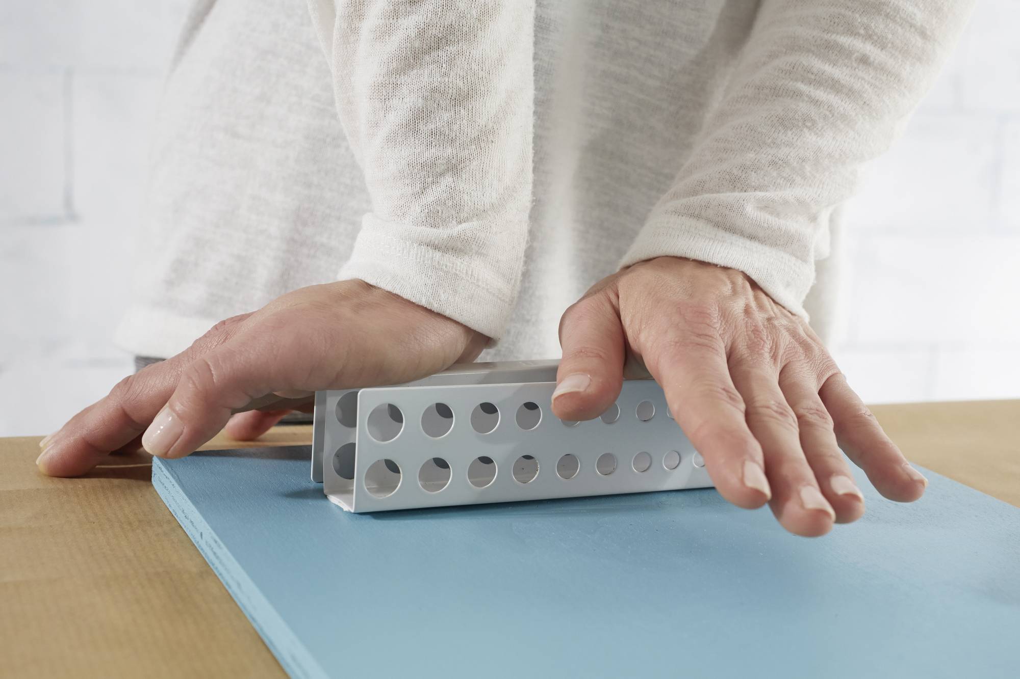 Hands are applying pressure to a metal template for woodworking to drill holes in a blue wooden board.