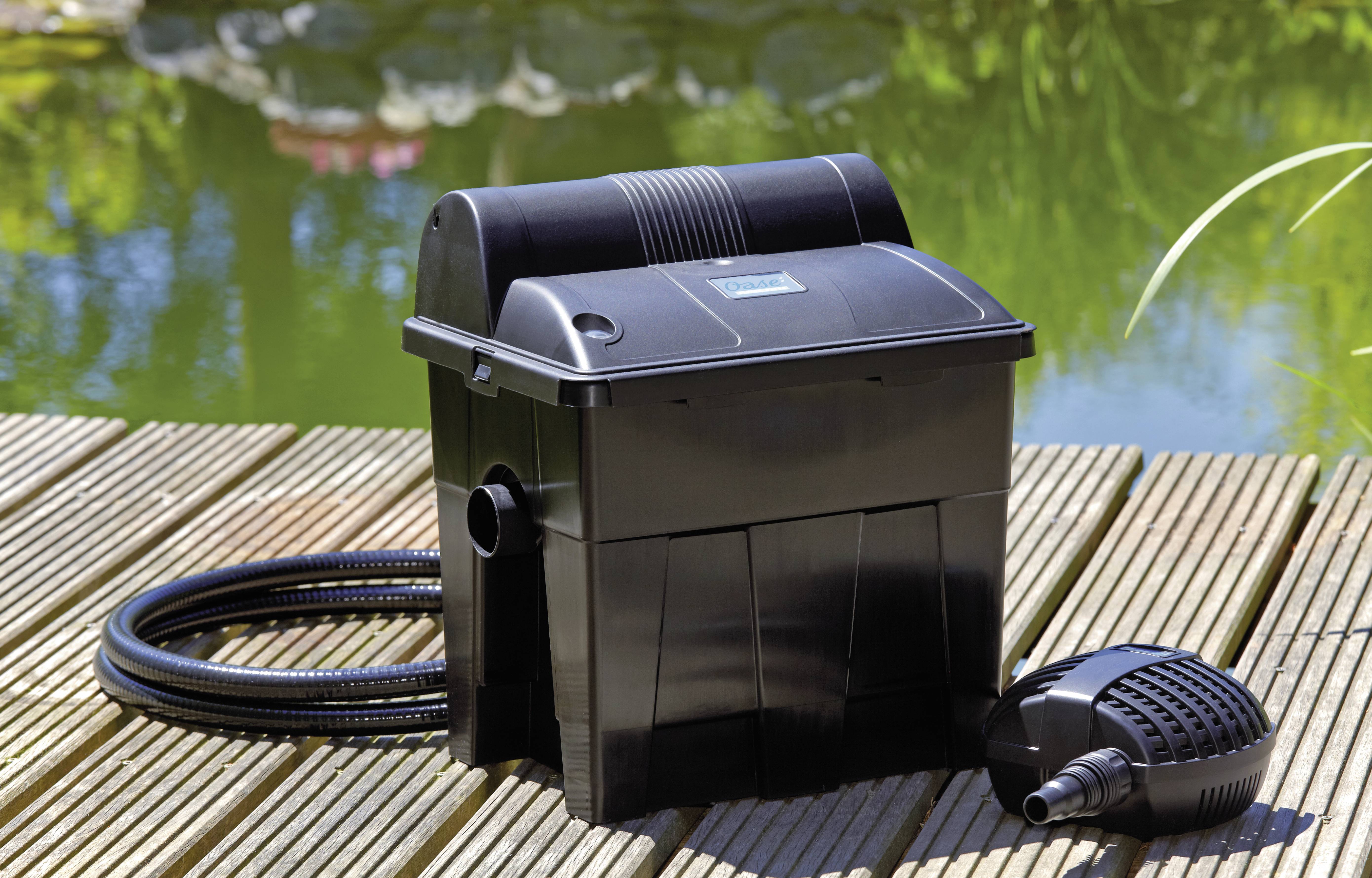 A black pond filter with a hose and pump on a wooden deck. A pond with greenery is in the background, suggesting a garden setting.