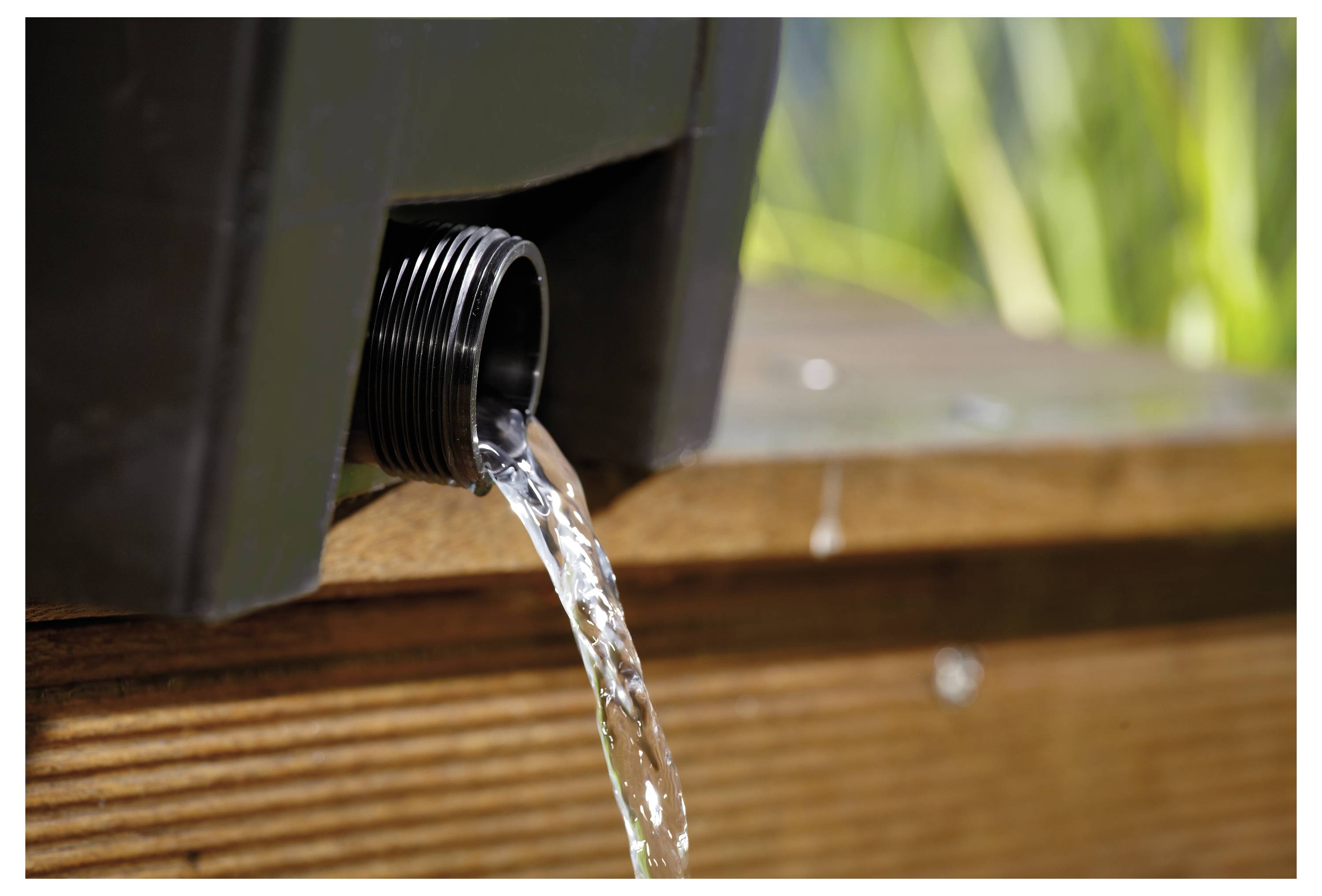 Water flows from a spout at the bottom of a dark container, demonstrating controlled release onto a wooden surface outdoors.