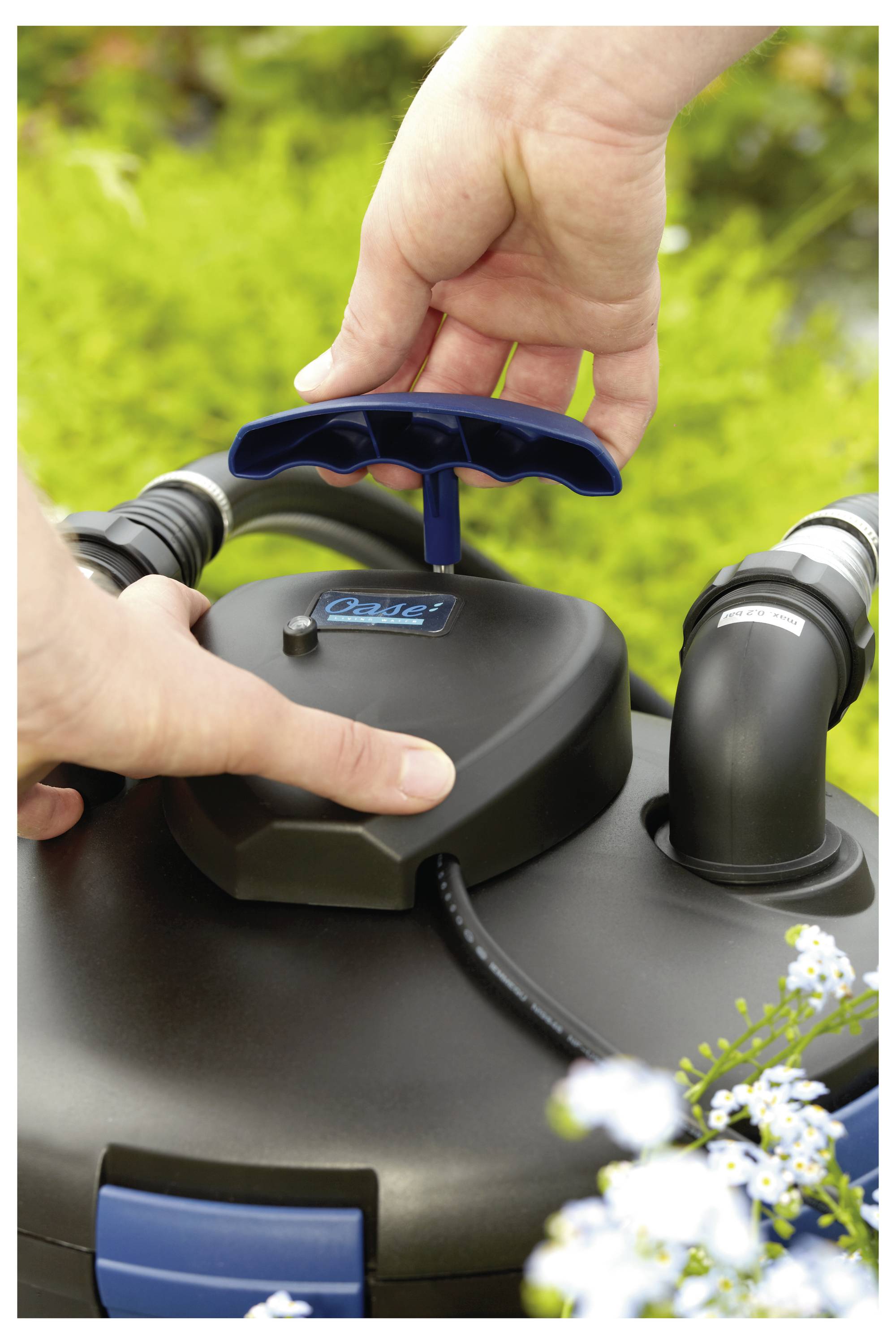 A person adjusts the handle of a black device with blue accents outdoors, surrounded by greenery and small flowers.