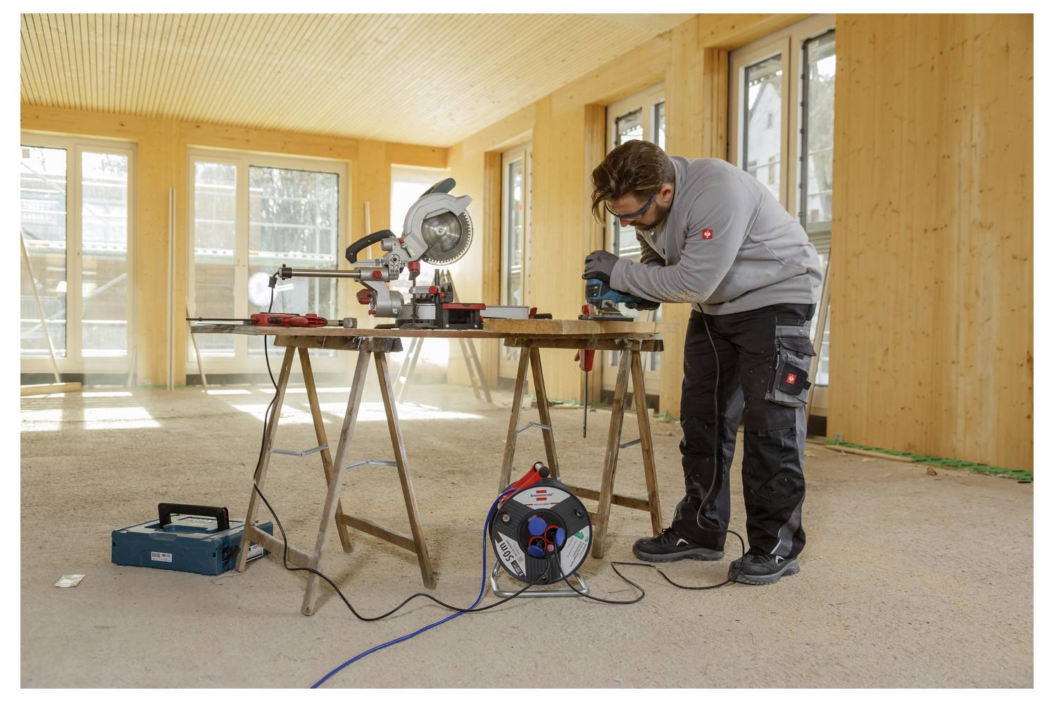 A person is using a saw on a wooden board in a partially constructed room, with windows and raw wooden walls visible in the background.