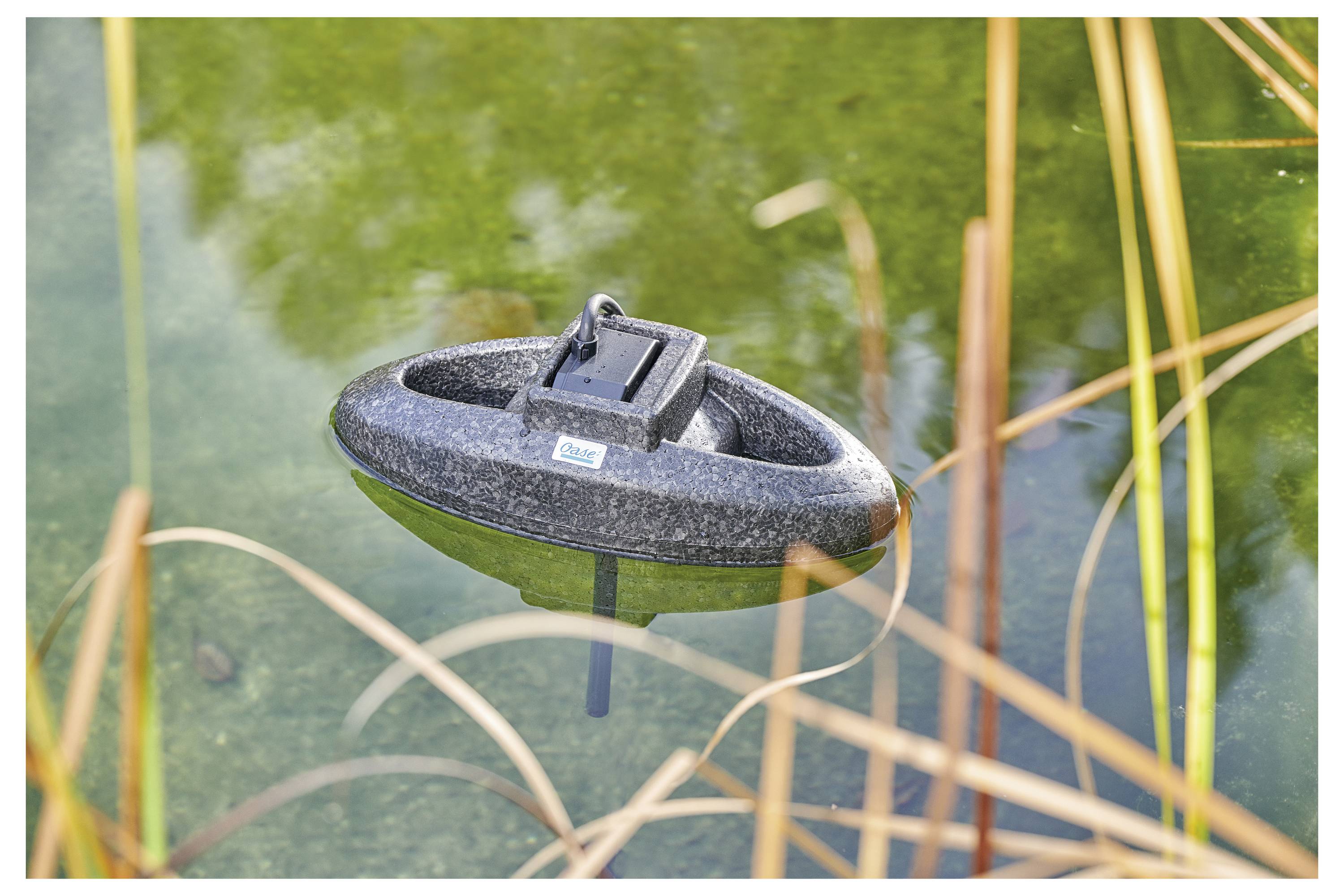 A floating black device in a pond, surrounded by reeds. It is used for monitoring water quality, with faint reflections on the water surface.