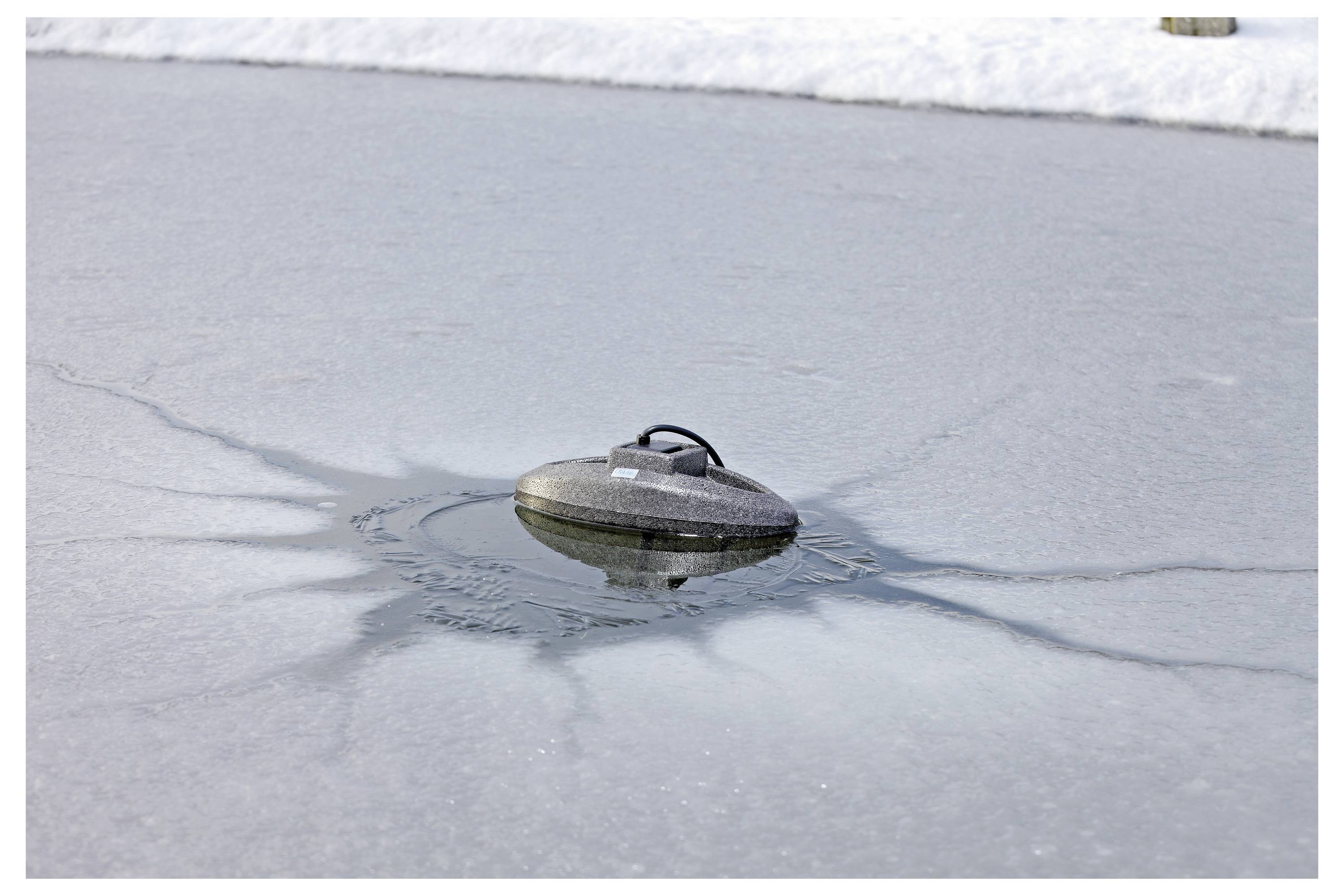 A small, floating gray object is on an icy pond, creating a melted circular area around it. Snow surrounds the pond in the background.