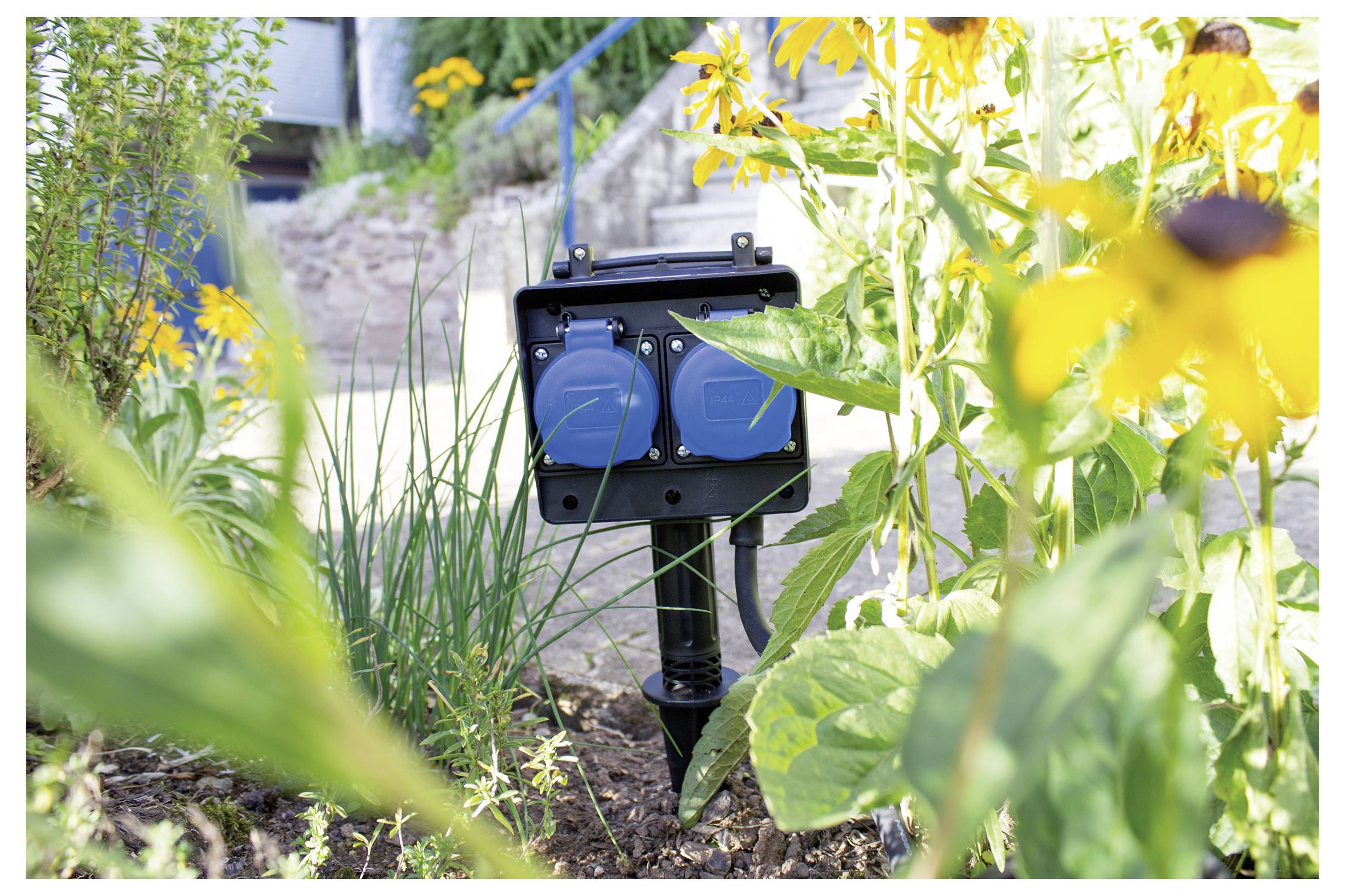 A black outdoor electrical outlet box with two covered sockets is surrounded by yellow flowers and green plants in a garden setting.