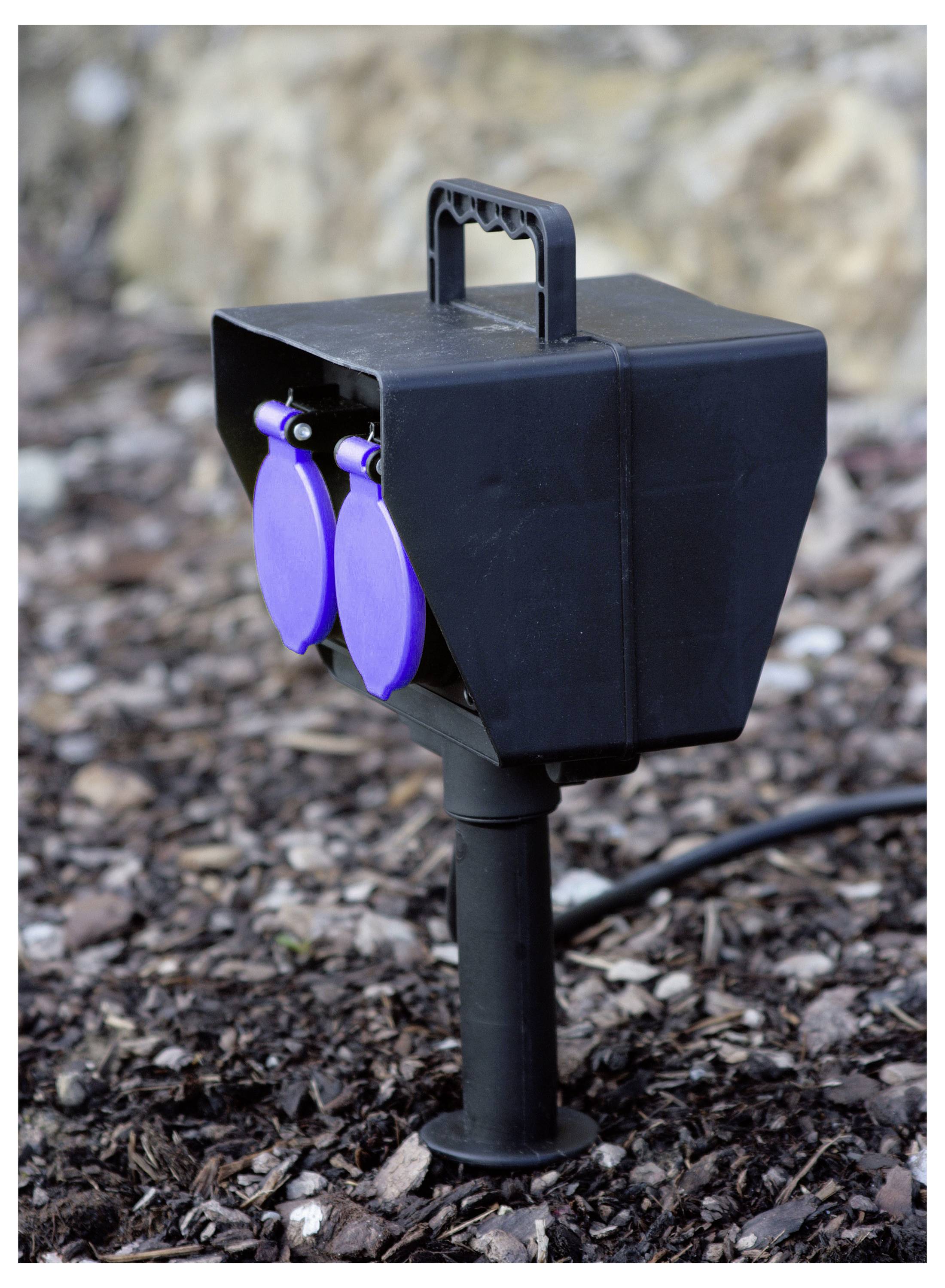 A black outdoor power outlet with two blue sockets is mounted on a post in a garden setting, surrounded by bark mulch.