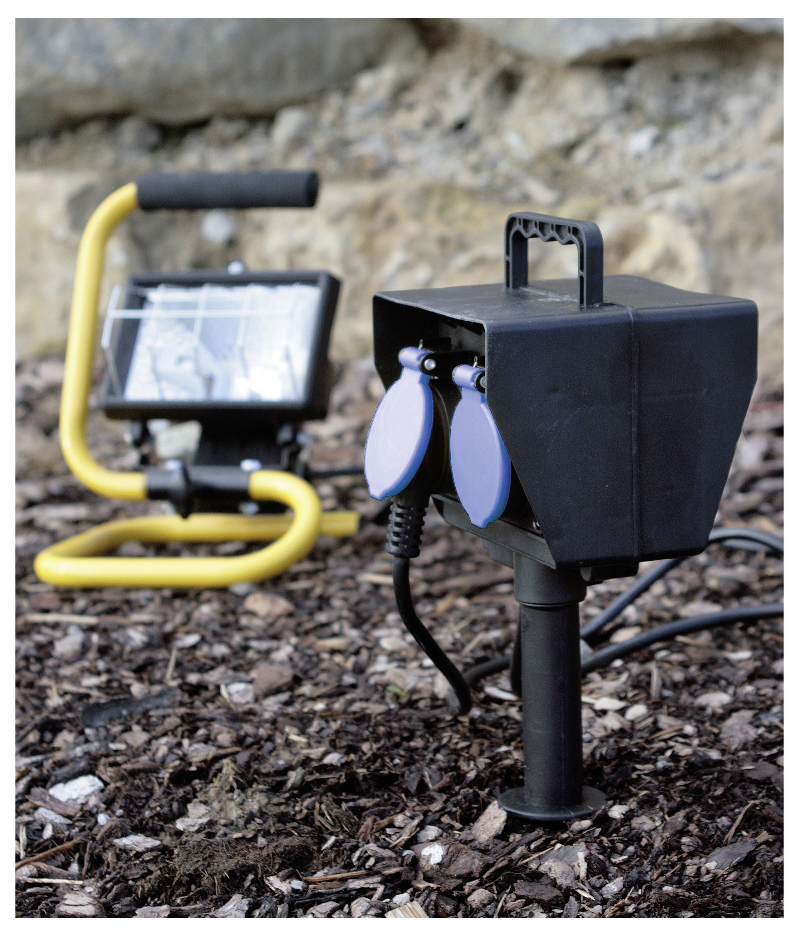 A solar-powered outdoor floodlight with a black and blue electrical outlet is staked into the ground in a garden setting.