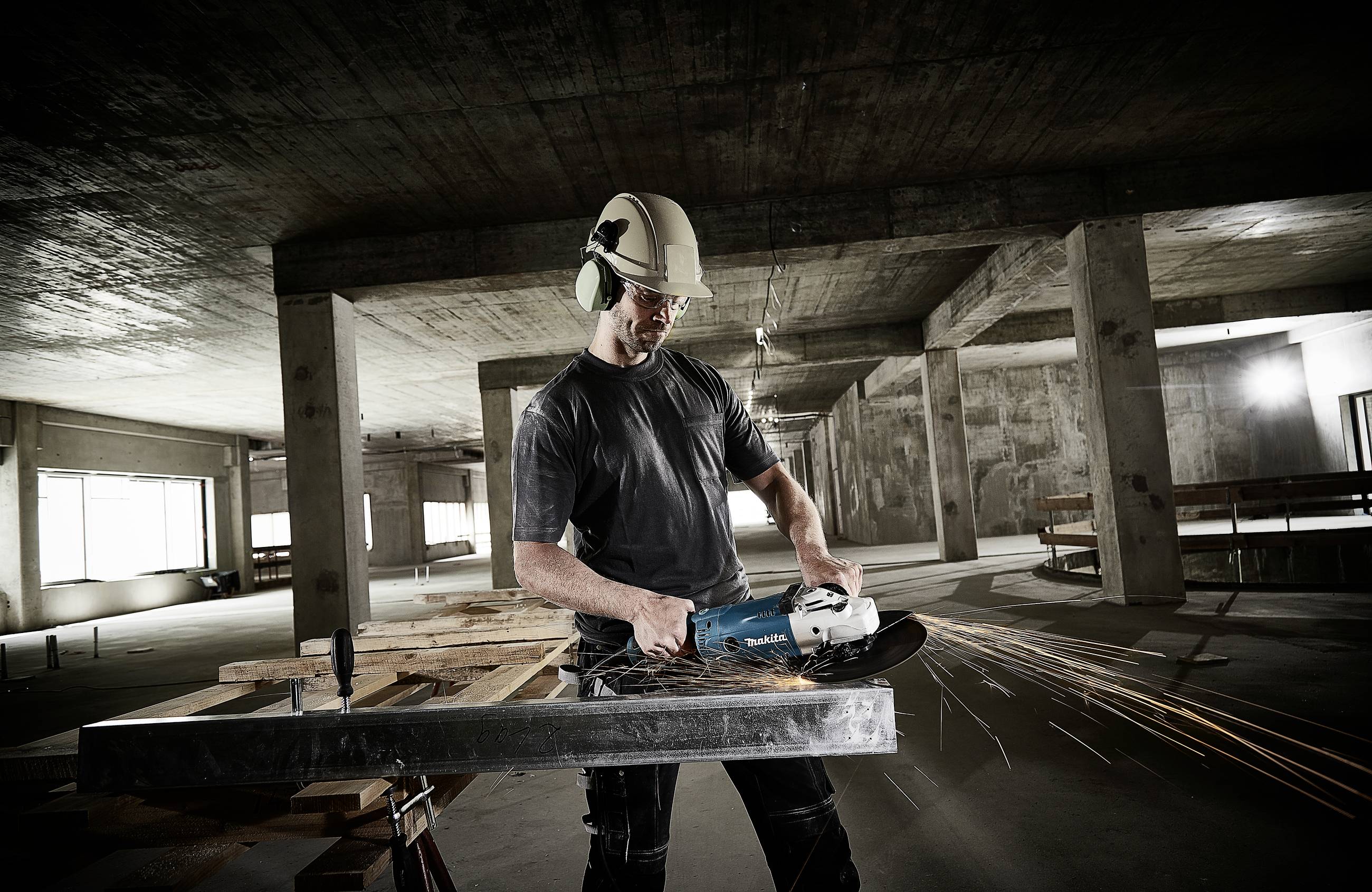 A construction worker wearing a hard hat and ear defenders is cutting a metal beam with an angle grinder on a building site. Sparks are flying.