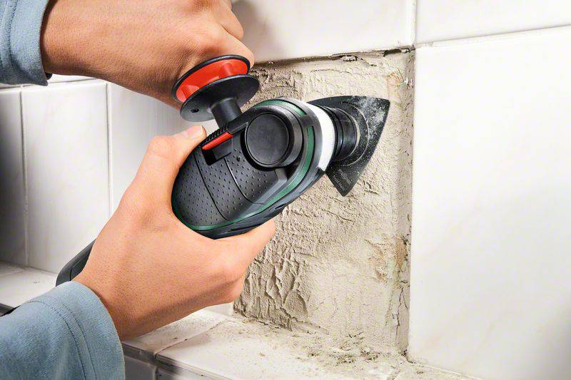 A person is using an electric tool to remove mortar between white tiles. The scene depicts craftsmanship at work.