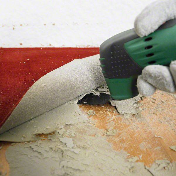 A person is removing a red carpet from a hard floor using an electric tool, with damage visible underneath the carpet.