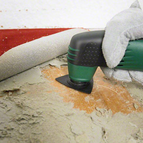 An electric sanding machine removes paint from a wooden surface. A hand wearing a grey glove firmly grips the tool.