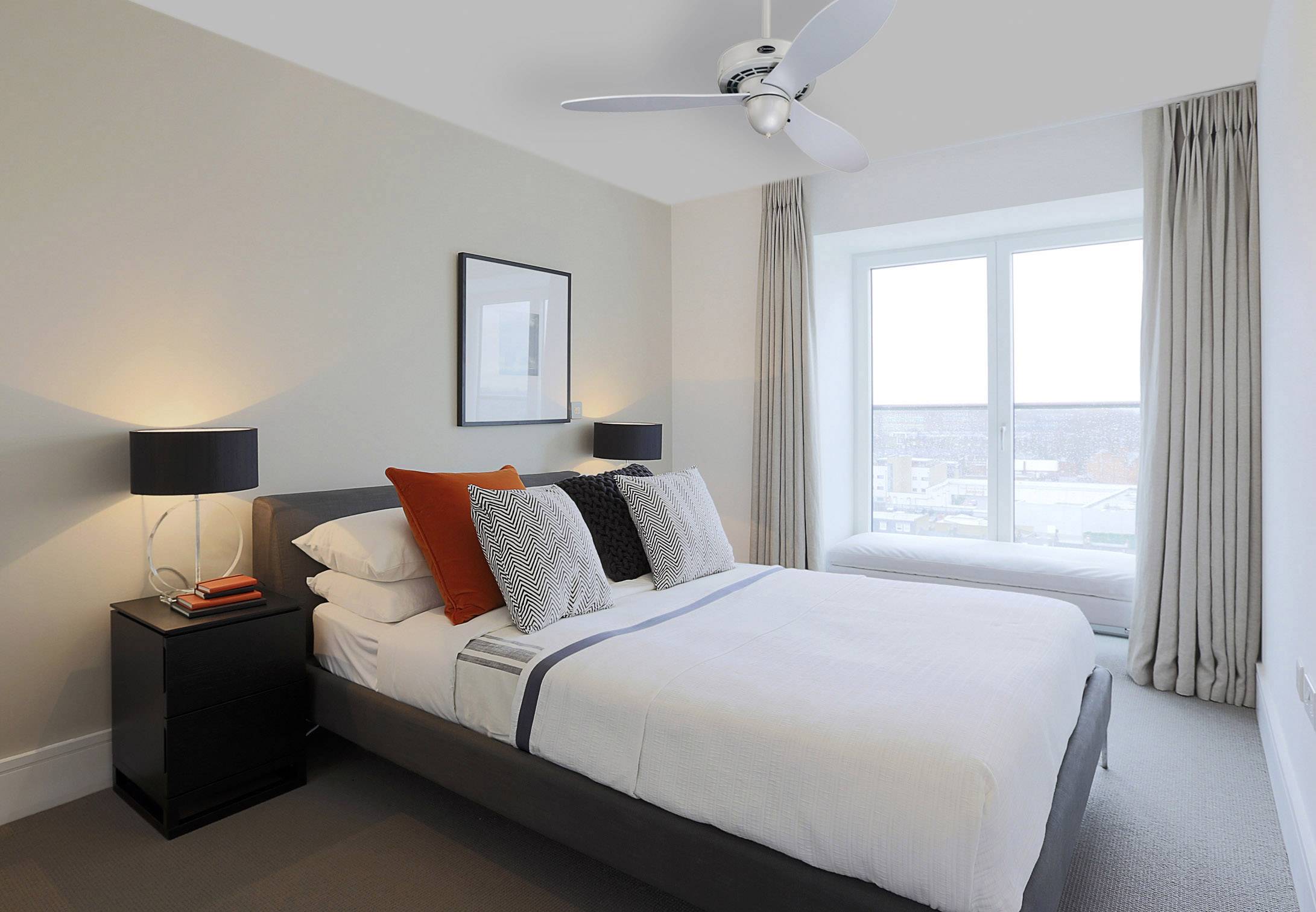 A bedroom with a double bed, decorated with orange and grey cushions. Two bedside tables with lamps, window in the background.