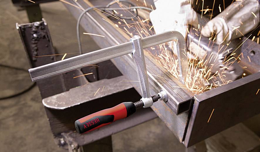 A G-clamp holds metal parts in place while a worker sprays sparks with an angle grinder. Processing in a workshop.