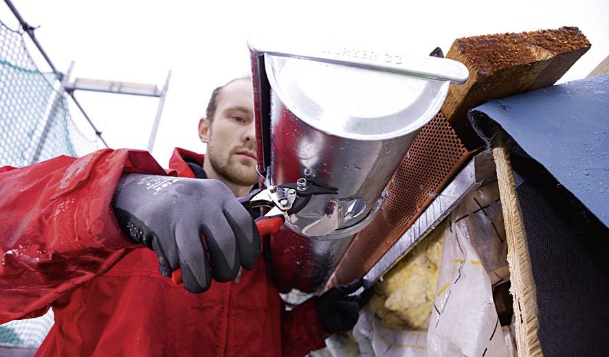 A person wearing a red jacket is installing a gutter with gloves and pliers. A scaffolding is visible in the background.