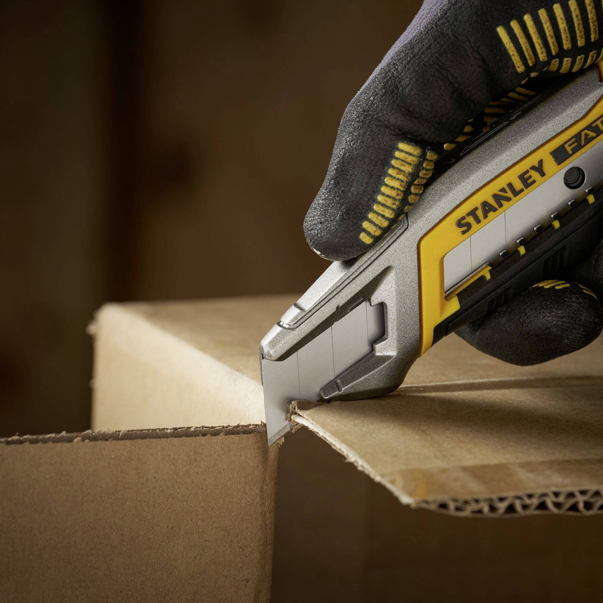 A gloved hand holds a craft knife, cutting cardboard.