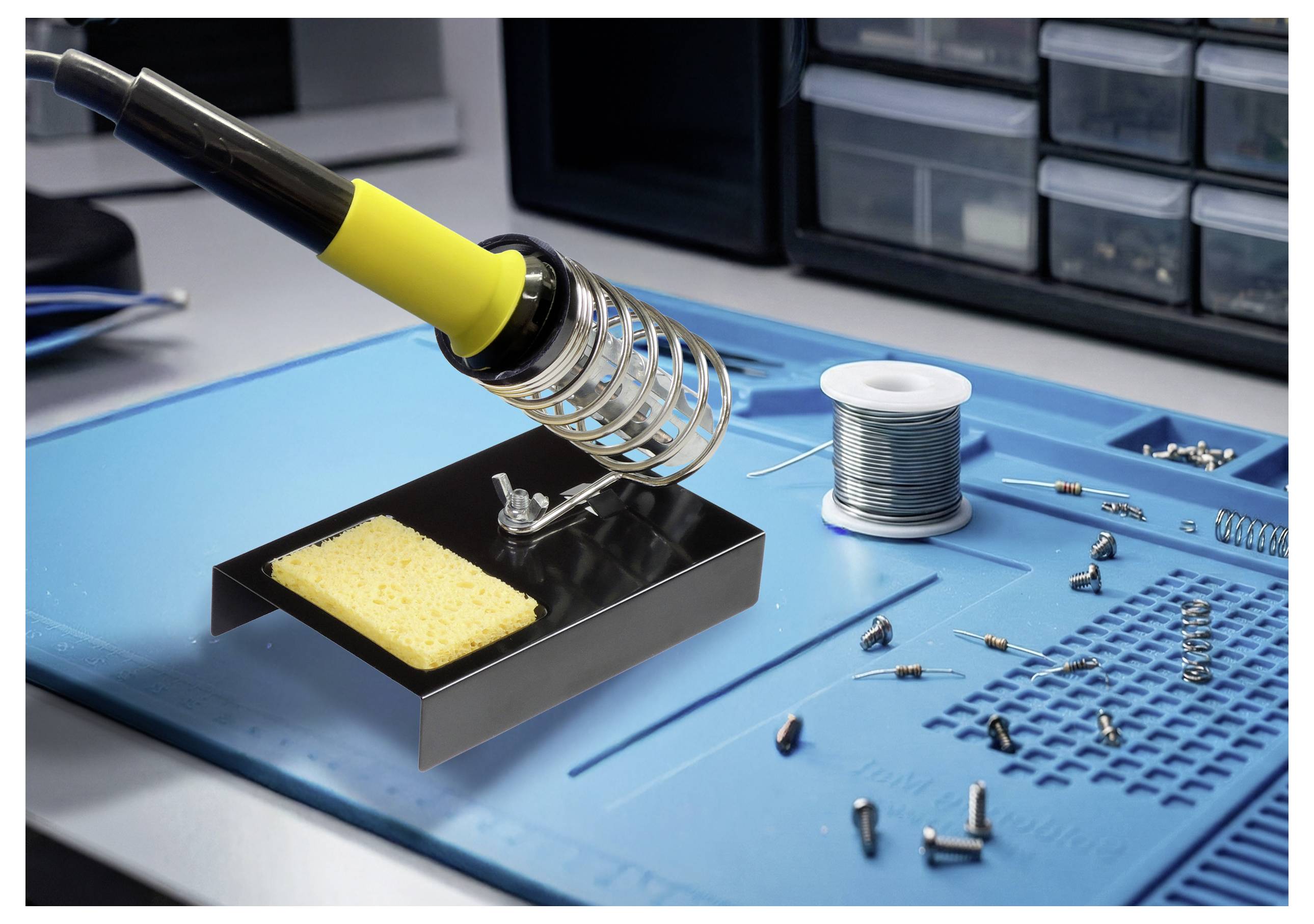 A soldering iron with a yellow handle rests in a stand on a blue mat. Nearby are a spool of solder and scattered electronic components.