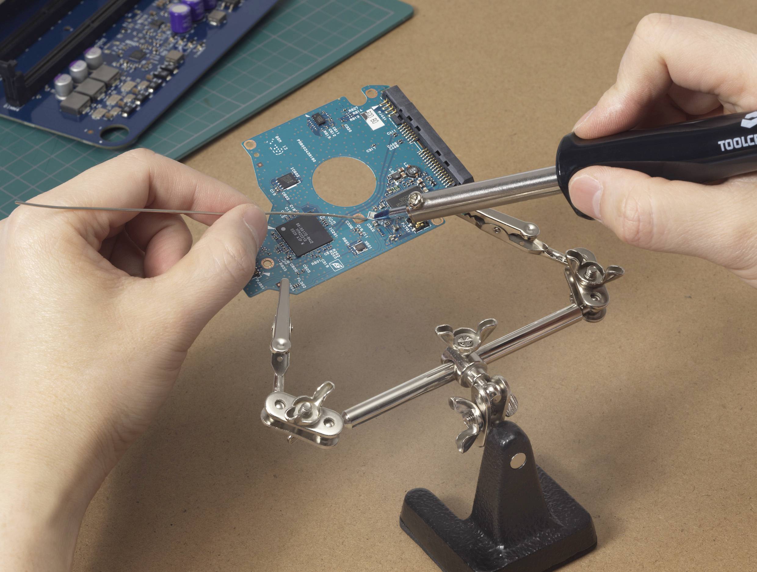 A person is soldering a circuit board with a soldering iron. The circuit board is being held in position by a clamp.