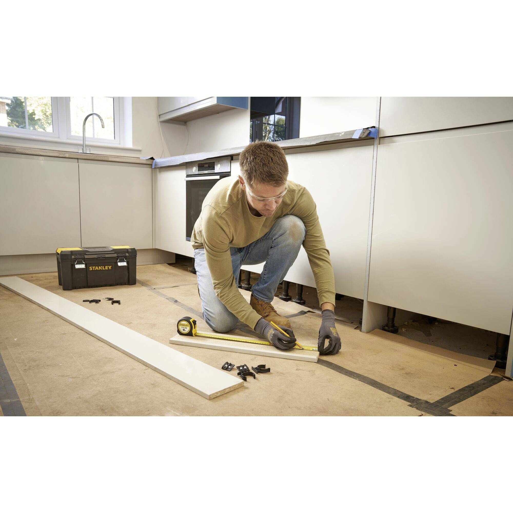 A person is measuring a wooden board on the floor of a kitchen. A toolbox is standing in the background.