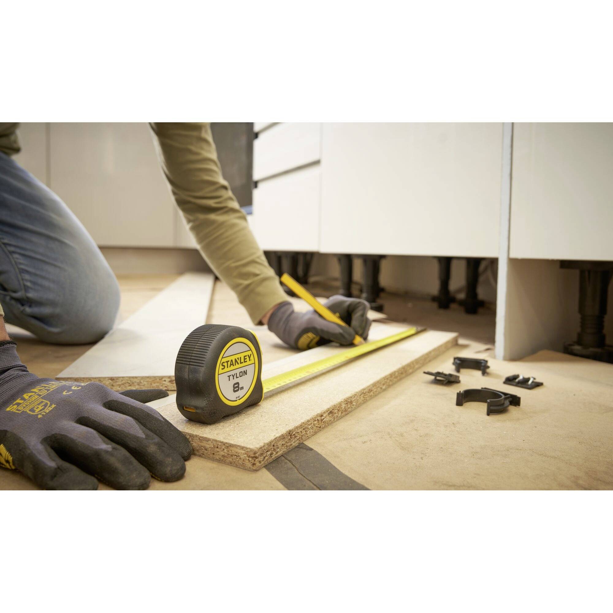 A person is measuring a wooden board with a tape measure and a pencil. Tools and cabinets are visible in the background.