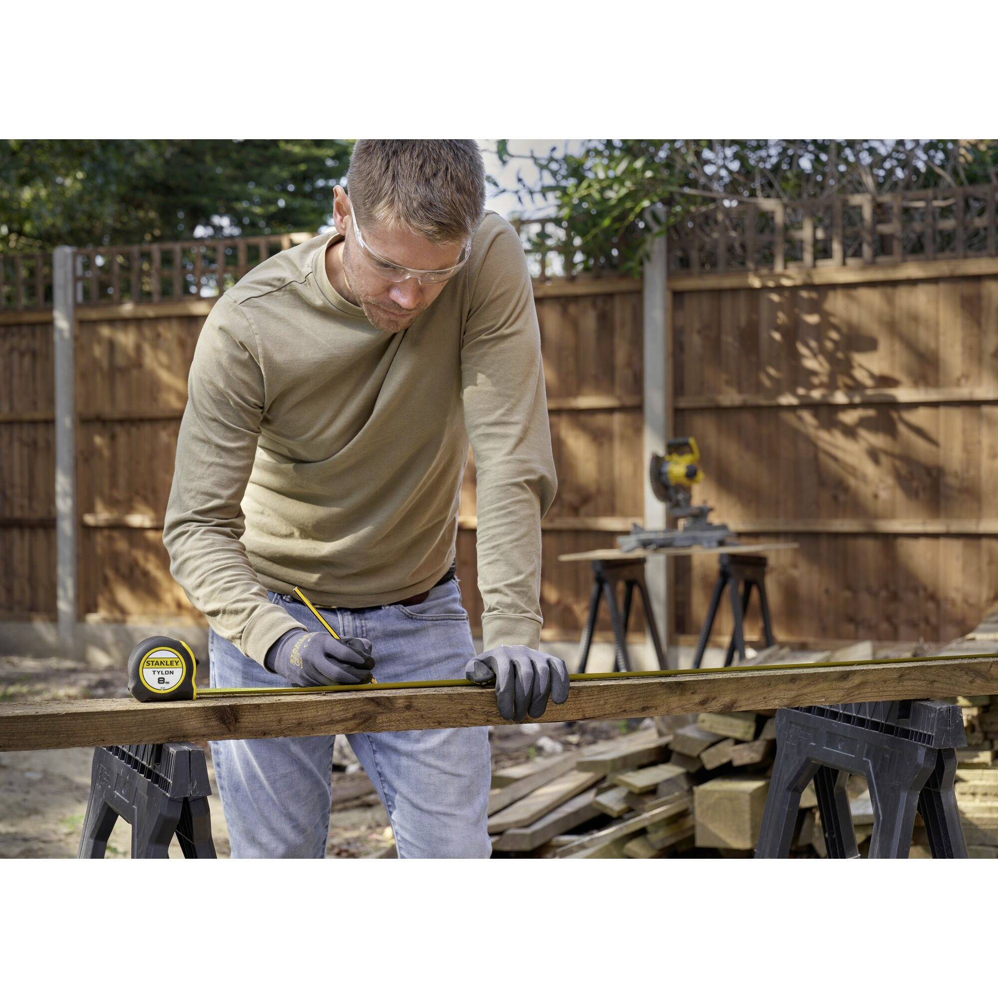 A person is wearing safety glasses and gloves and measuring a wooden board with a tape measure in the garden. A saw is visible in the background.