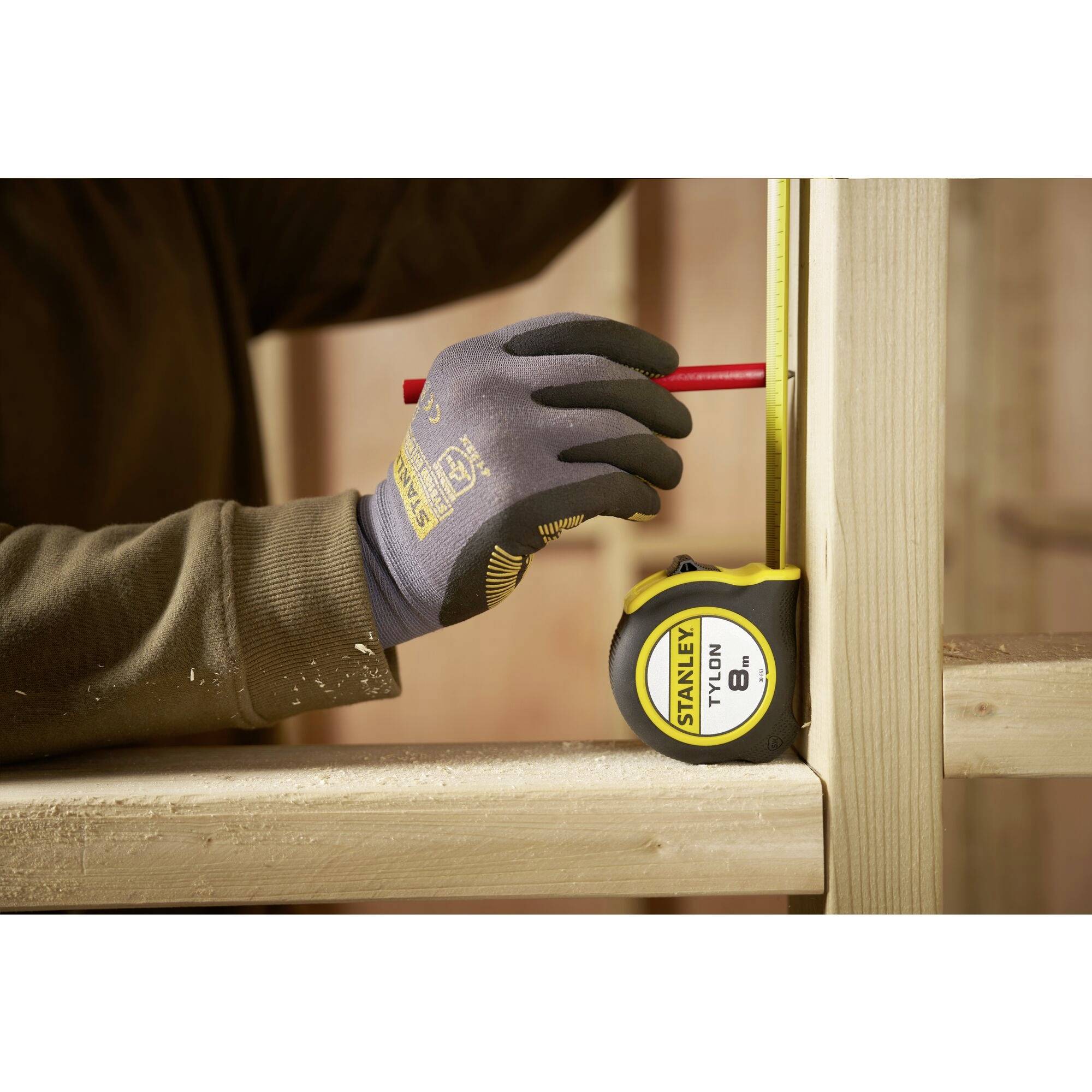 A person is measuring with a tape measure and marking with a pencil on a wooden frame. They are wearing a glove.