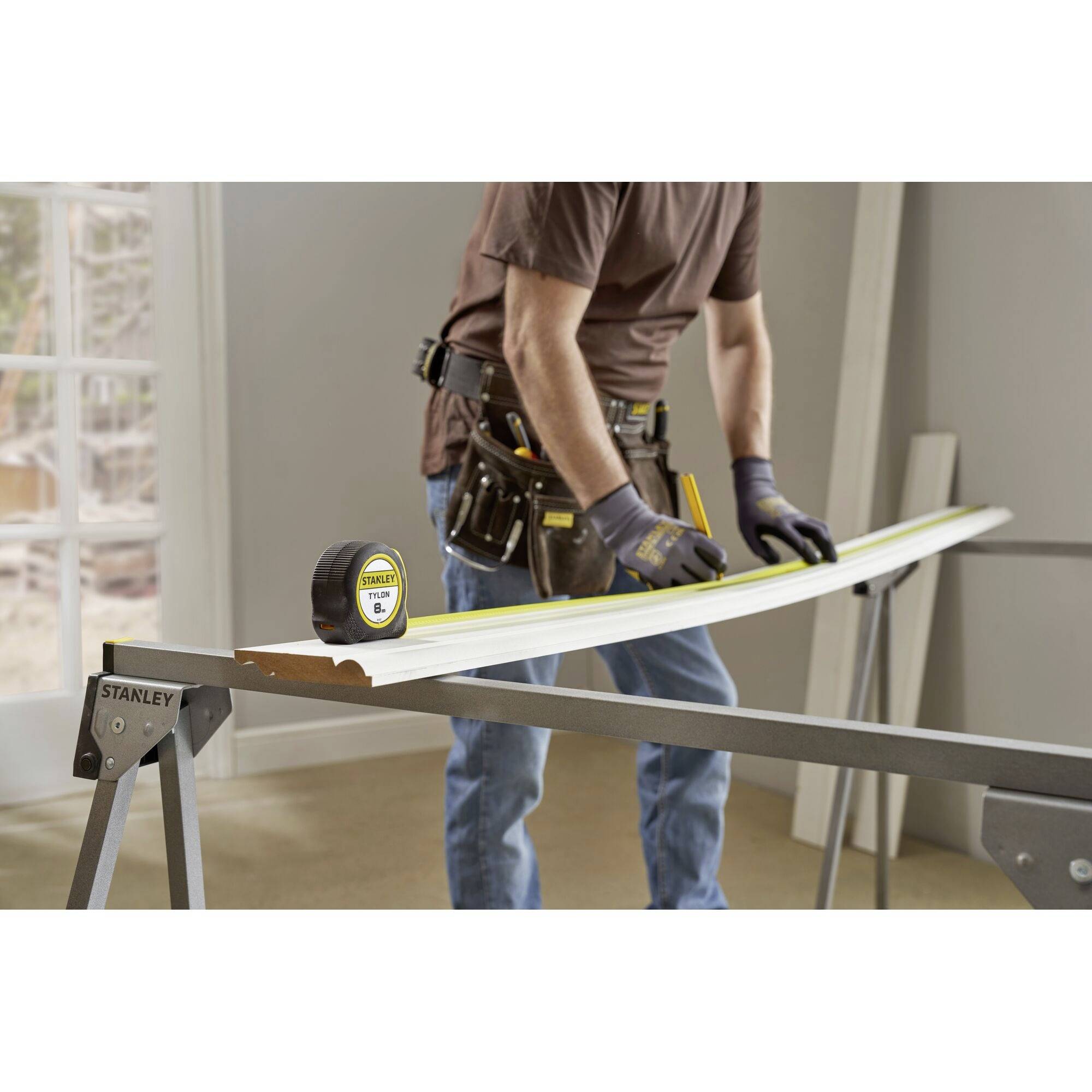 A craftsman is measuring a long plank on a sawbench using a tape measure. He is wearing gloves and carrying a tool belt.