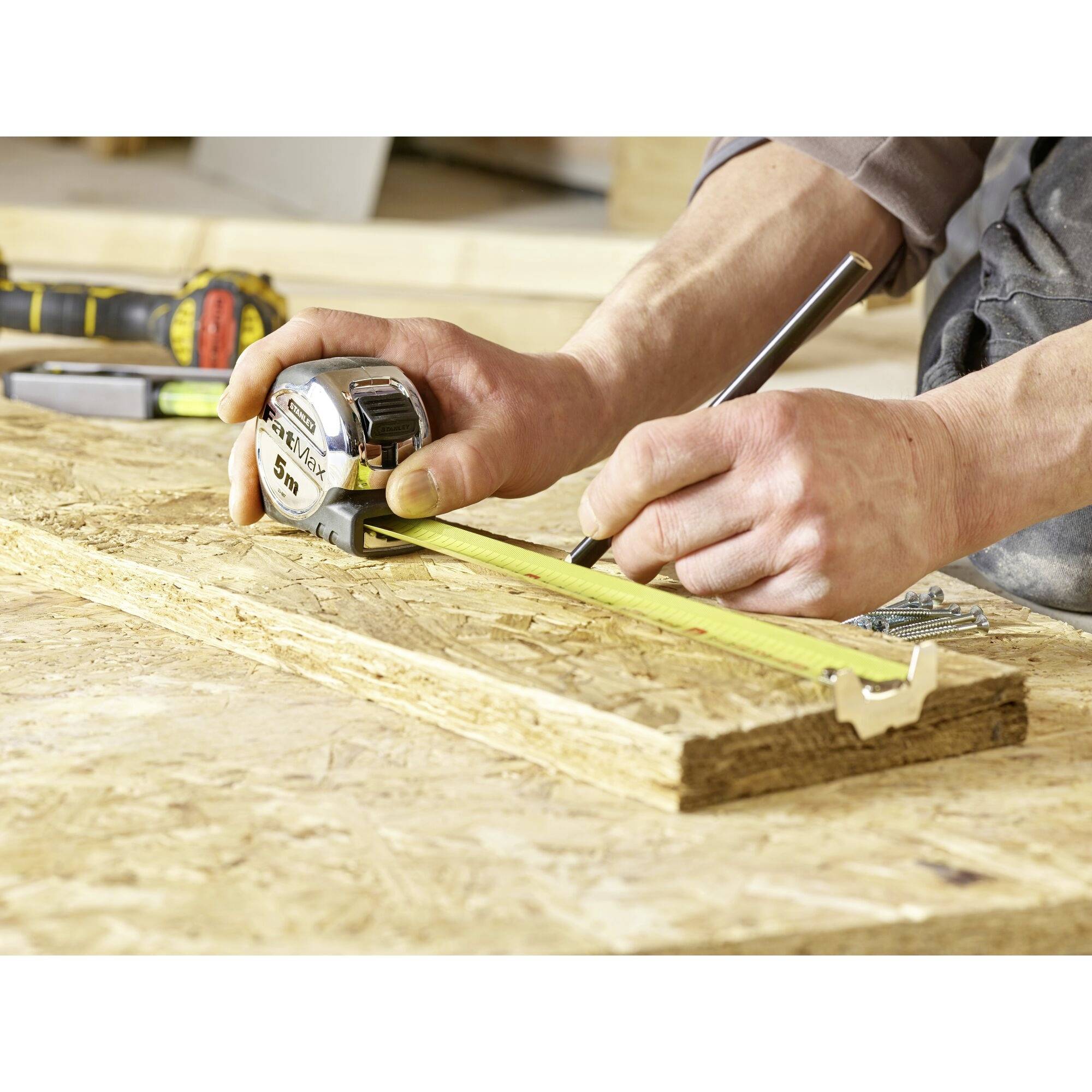 A person is measuring a wooden board with a tape measure and marking it with a pencil. An electric power tool can be seen in the background.