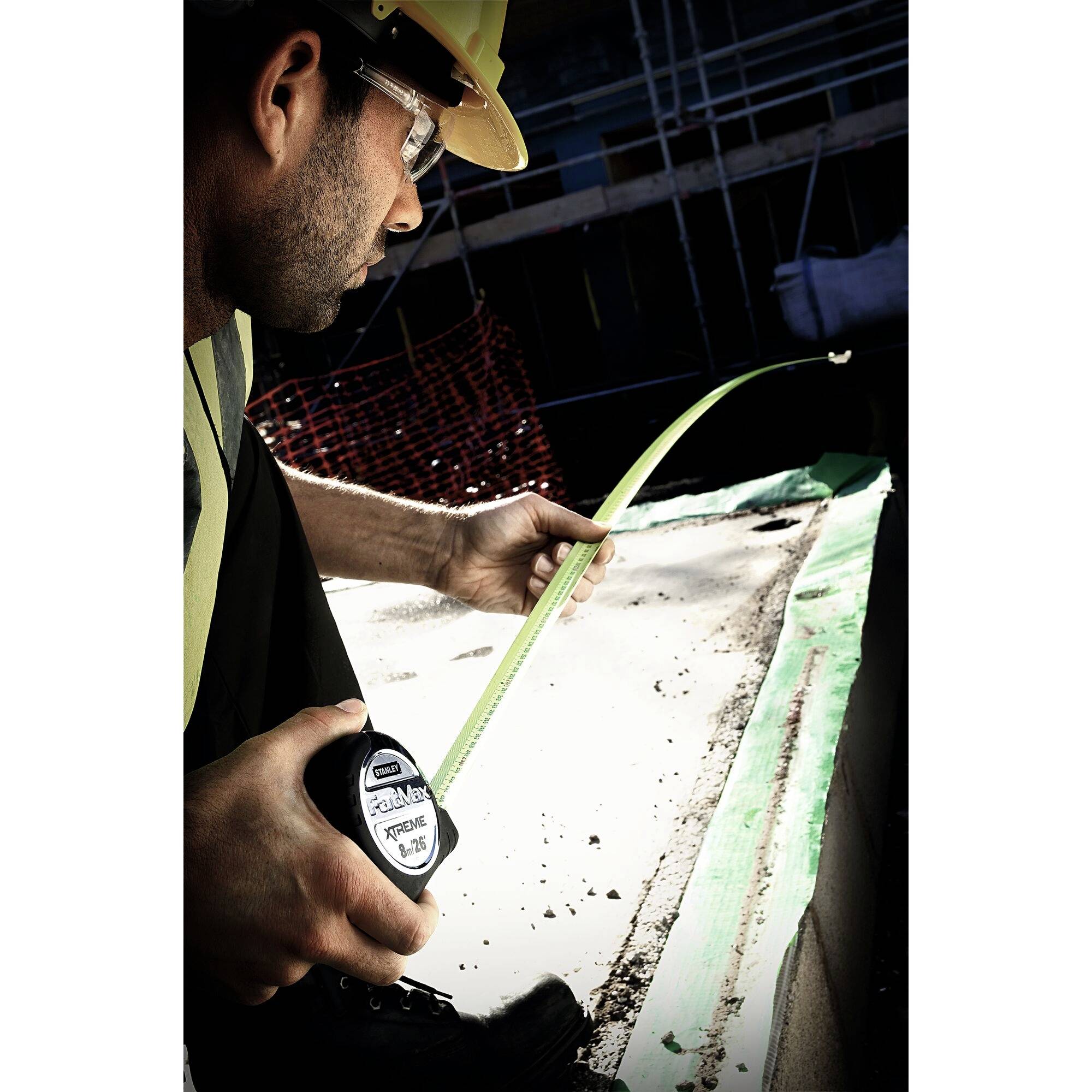 A construction worker wearing a hard hat and safety glasses is measuring a piece of wood with a tape measure on a building site.