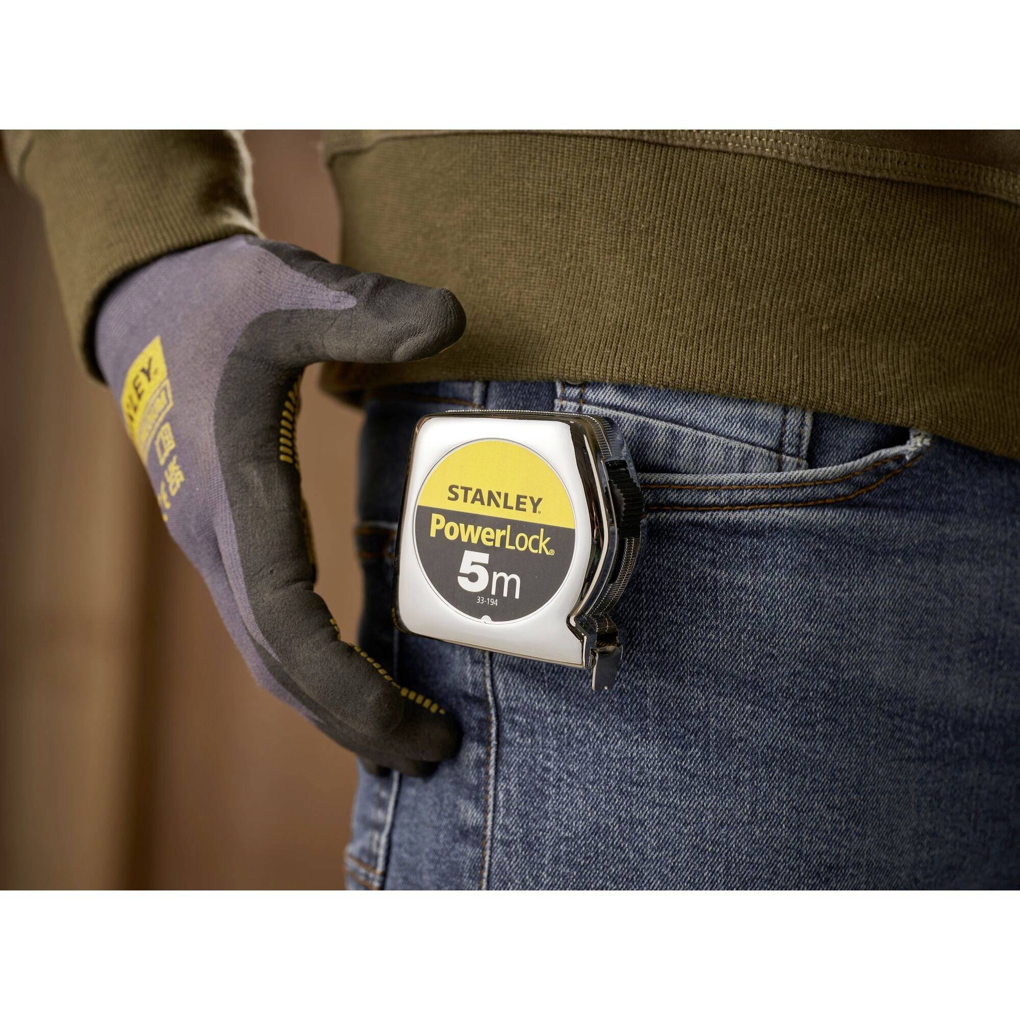 A person is wearing a 'Stanley PowerLock 5m' measuring tape on their belt, holding it with a gloved hand. Background is blurred, with focus on the measuring tape.