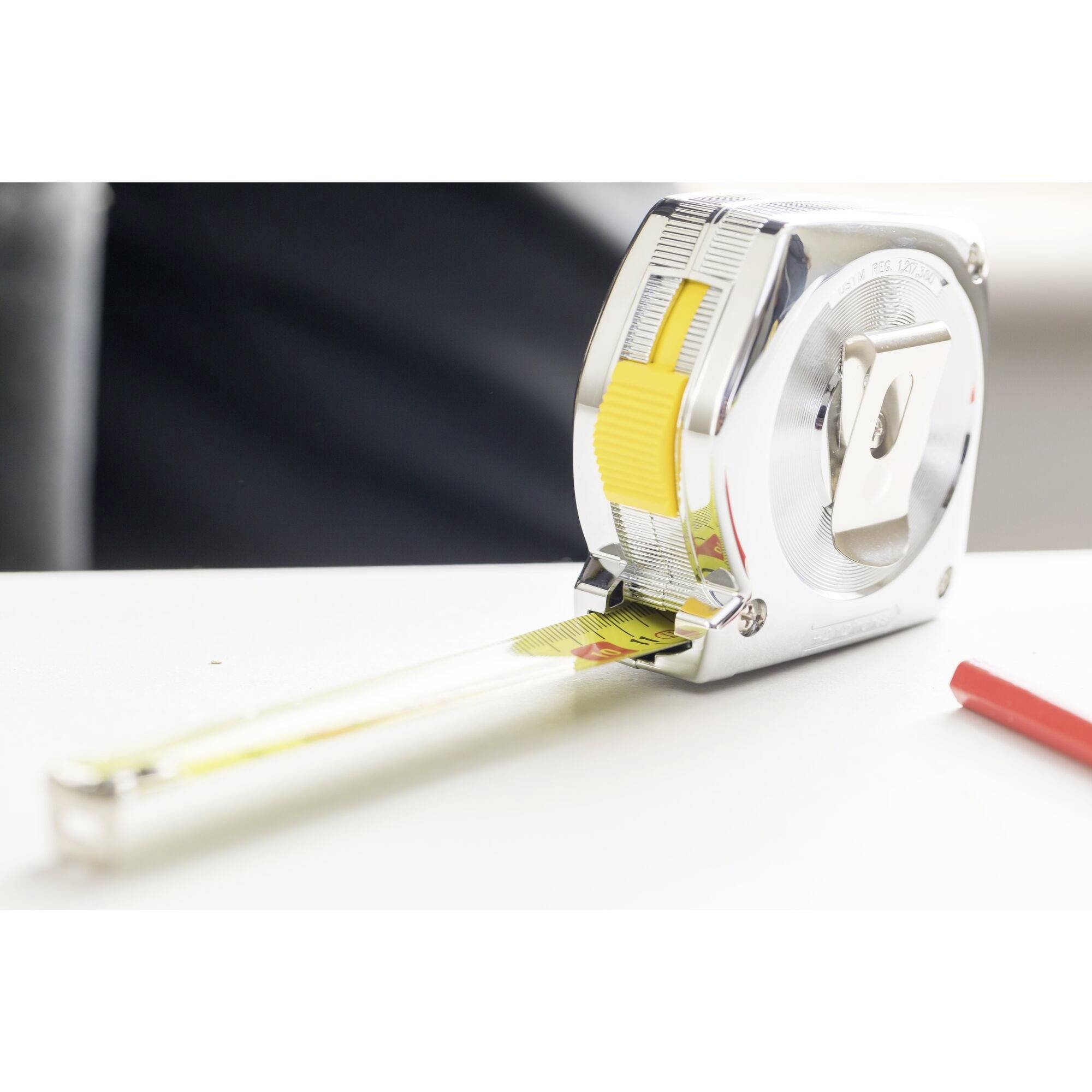 A silver tape measure lies on a table, with the tape partially extended. A red pen is beside it.