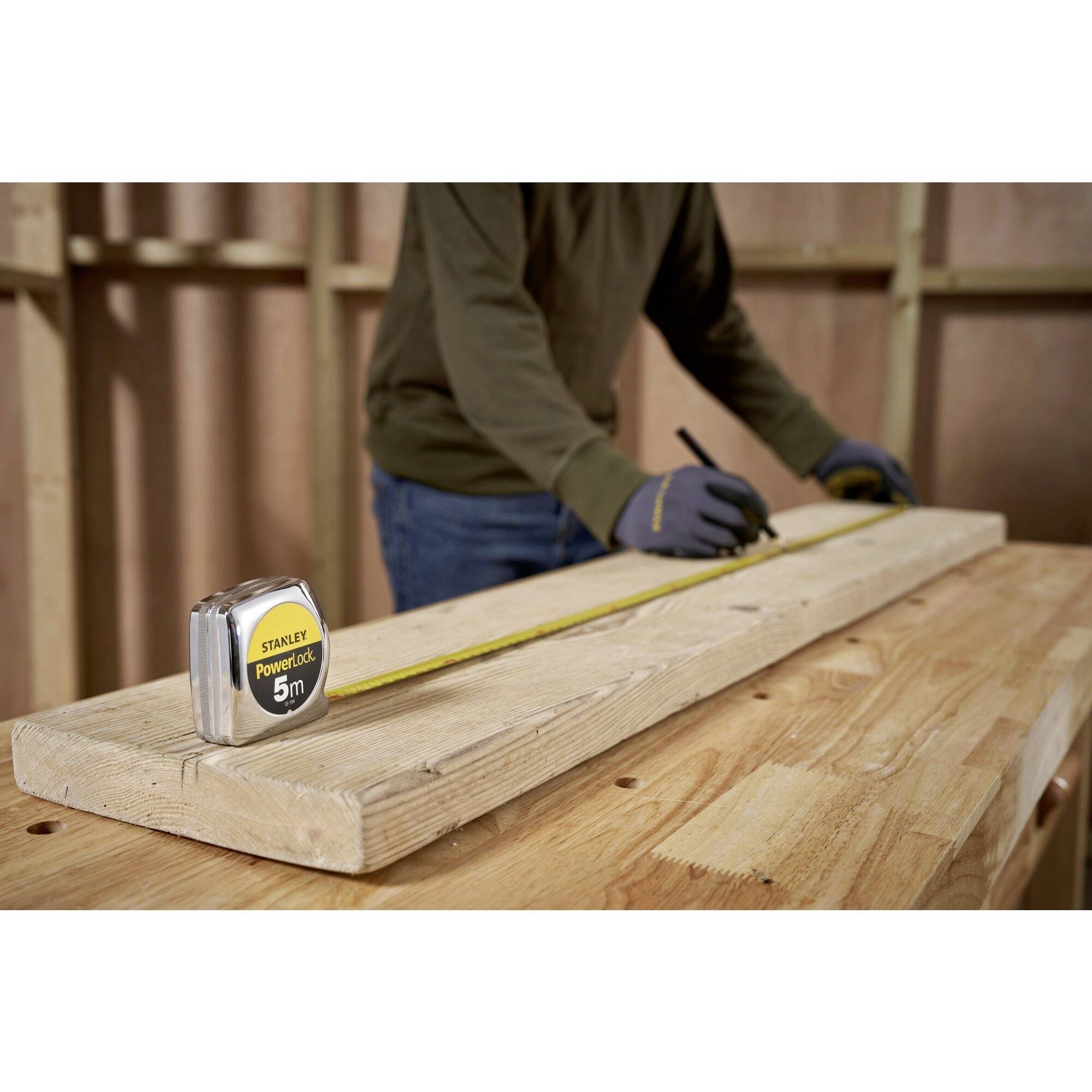 A person is measuring and marking a piece of wood in a workshop using a 5m measuring tape.