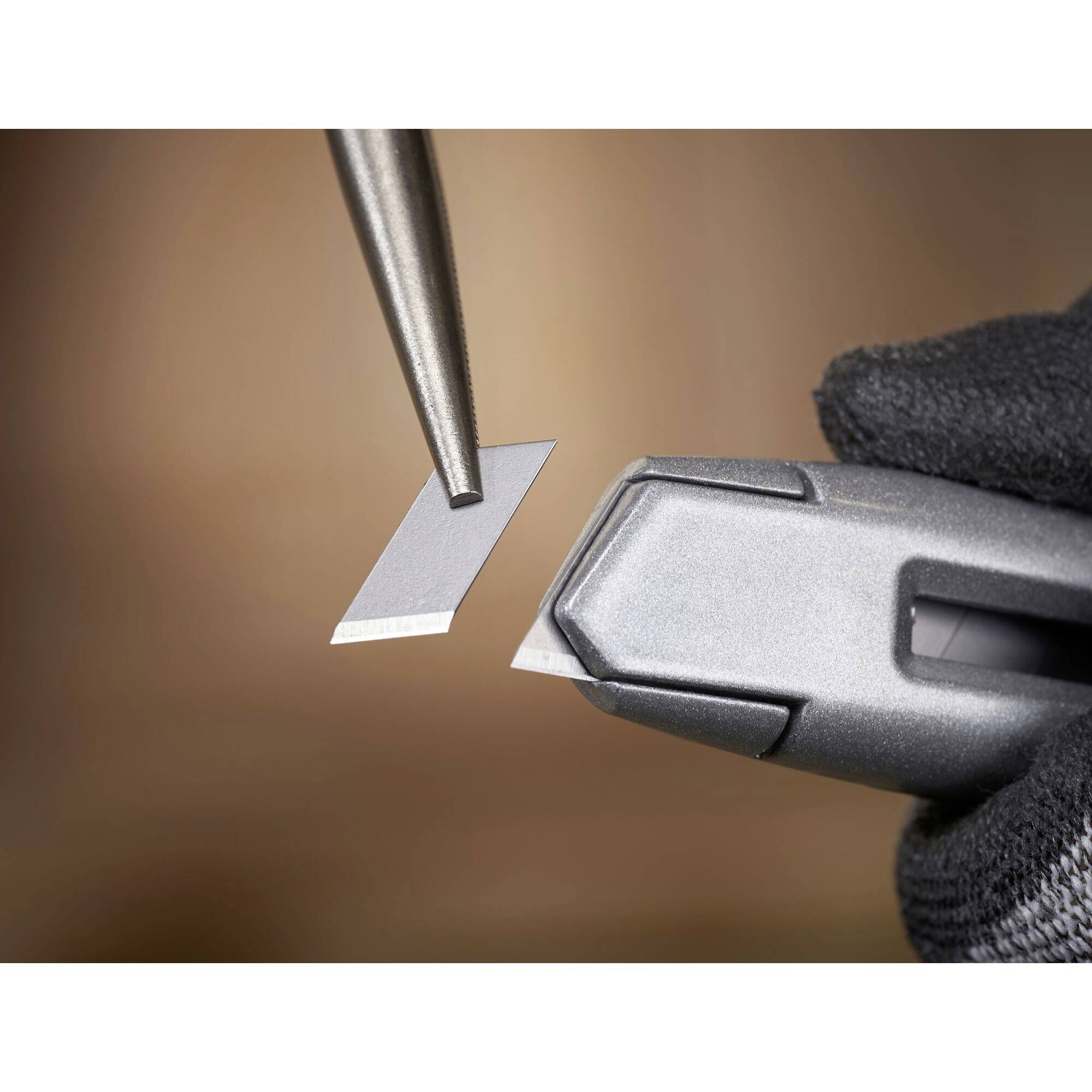 A person wearing a glove is holding a grey workpiece while touching a small, sharp-edged blade with tweezers.