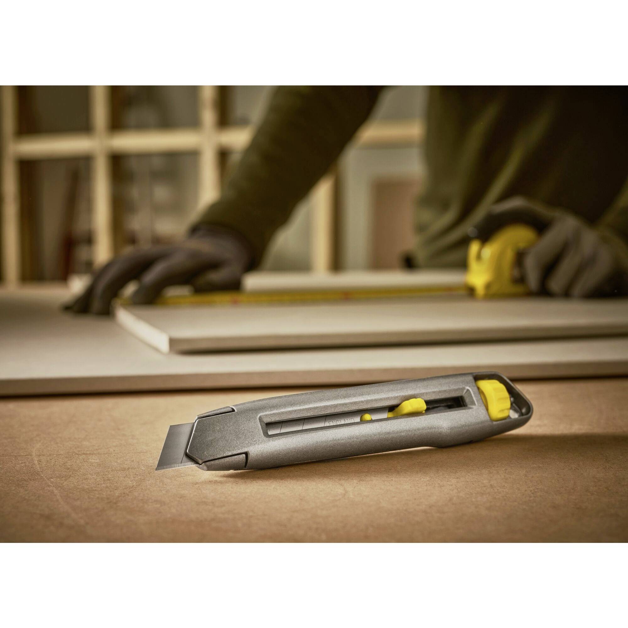 A person is measuring a wooden board with a tape measure, with a craft knife lying in the foreground. They are wearing gloves.