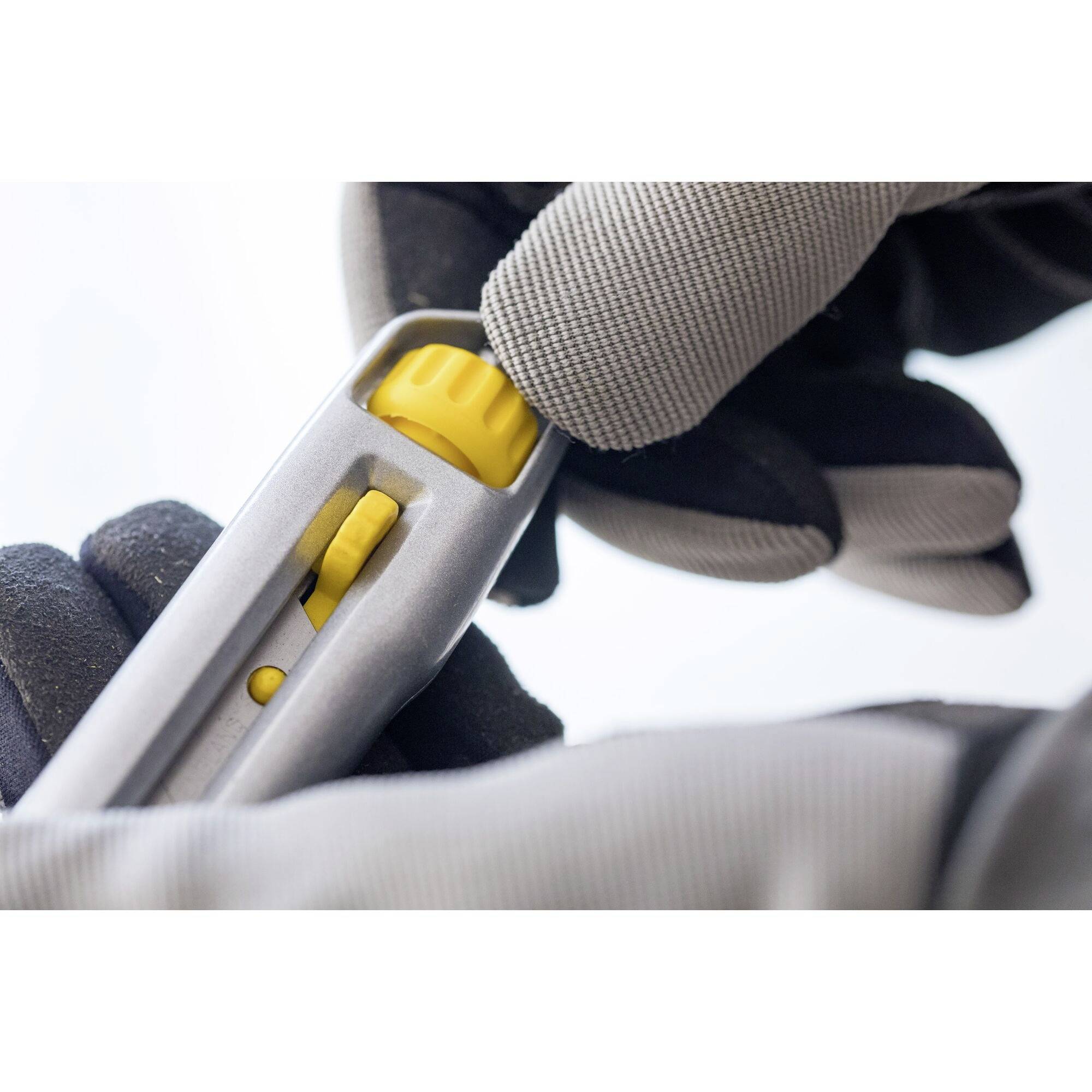 A gloved hand is holding a silver Stanley knife with a yellow blade and yellow adjustment wheel.