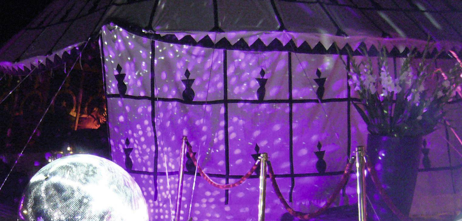 A festively illuminated scene with a white tent wall, purple light projections, and a large, gleaming disco ball in the foreground.
