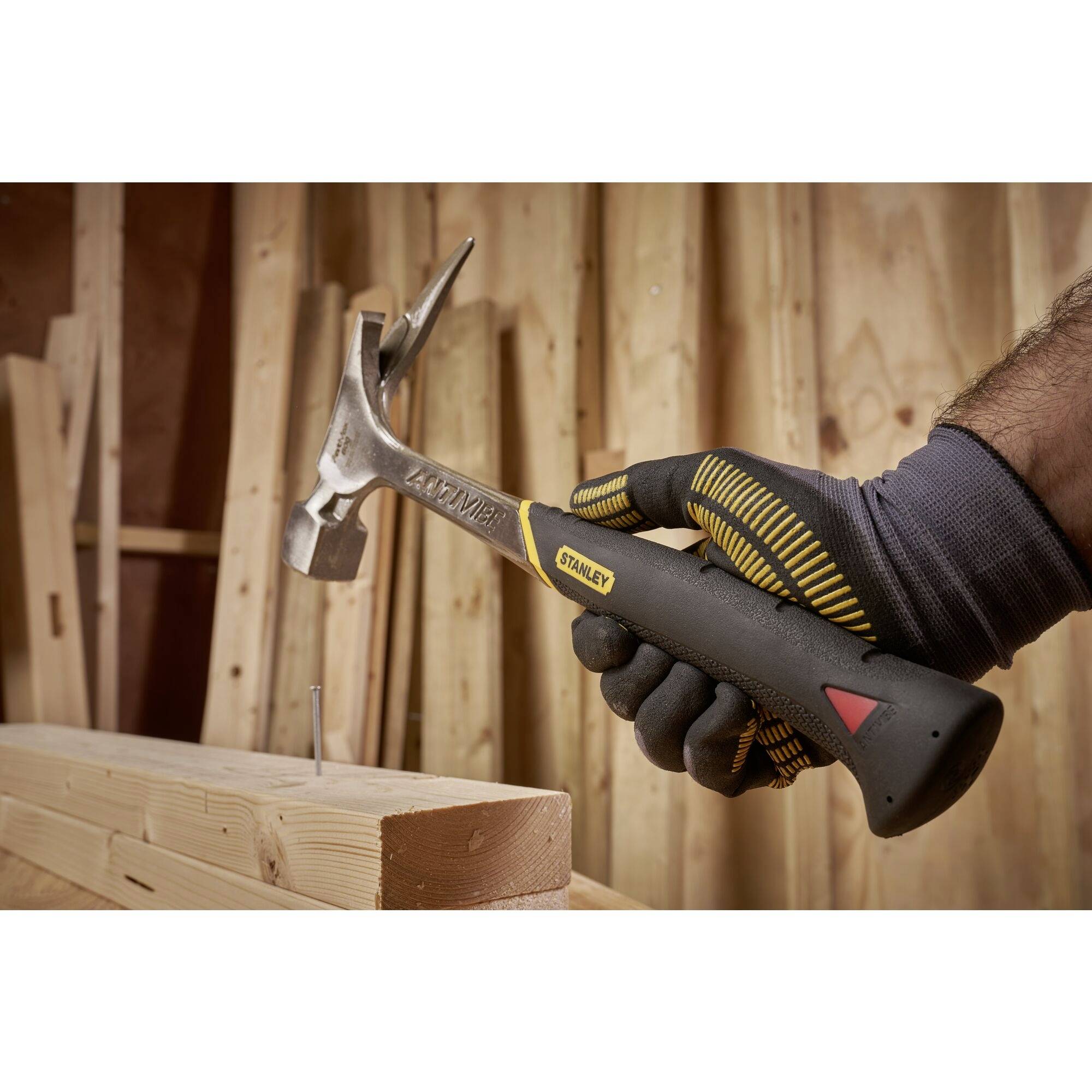 A hand grips a hammer with a yellow handle, driving a nail into a piece of wood in a workshop. The background consists of wooden laths.