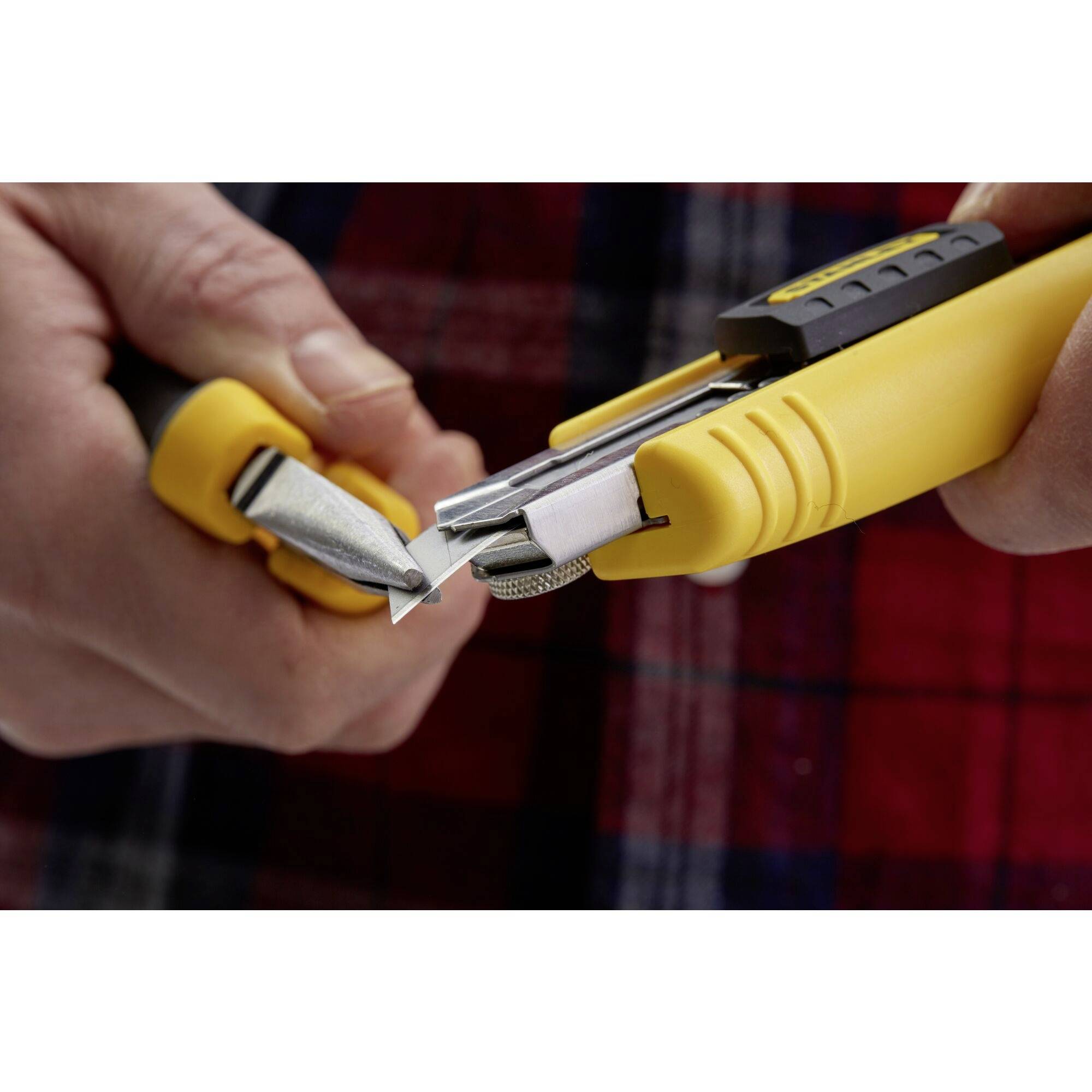 Two hands are holding a yellow utility knife and a replacement blade dispenser. A blade is being changed. Background: checked shirt.